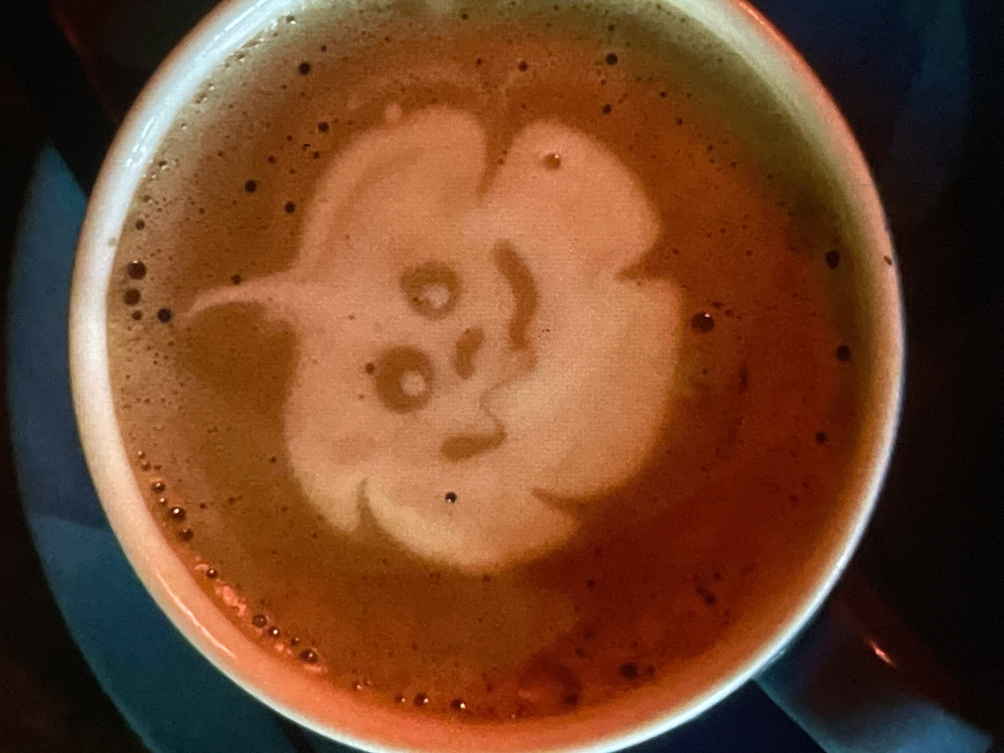 The image shows a close-up of a white coffee cup filled with latte art. The latte art depicts a jack-o'-lantern face, complete with two circular eyes, a triangular nose, and a smiling mouth. The foam art is created with a lighter-colored milk foam against the darker coffee. The cup is placed on a dark saucer, which is itself on a dark surface. A hint of a teal-colored object is visible in the upper left corner. The lighting is warm and somewhat dim, highlighting the rich colors of the coffee and foam. This image was taken in Harmanli, Bulgaria.