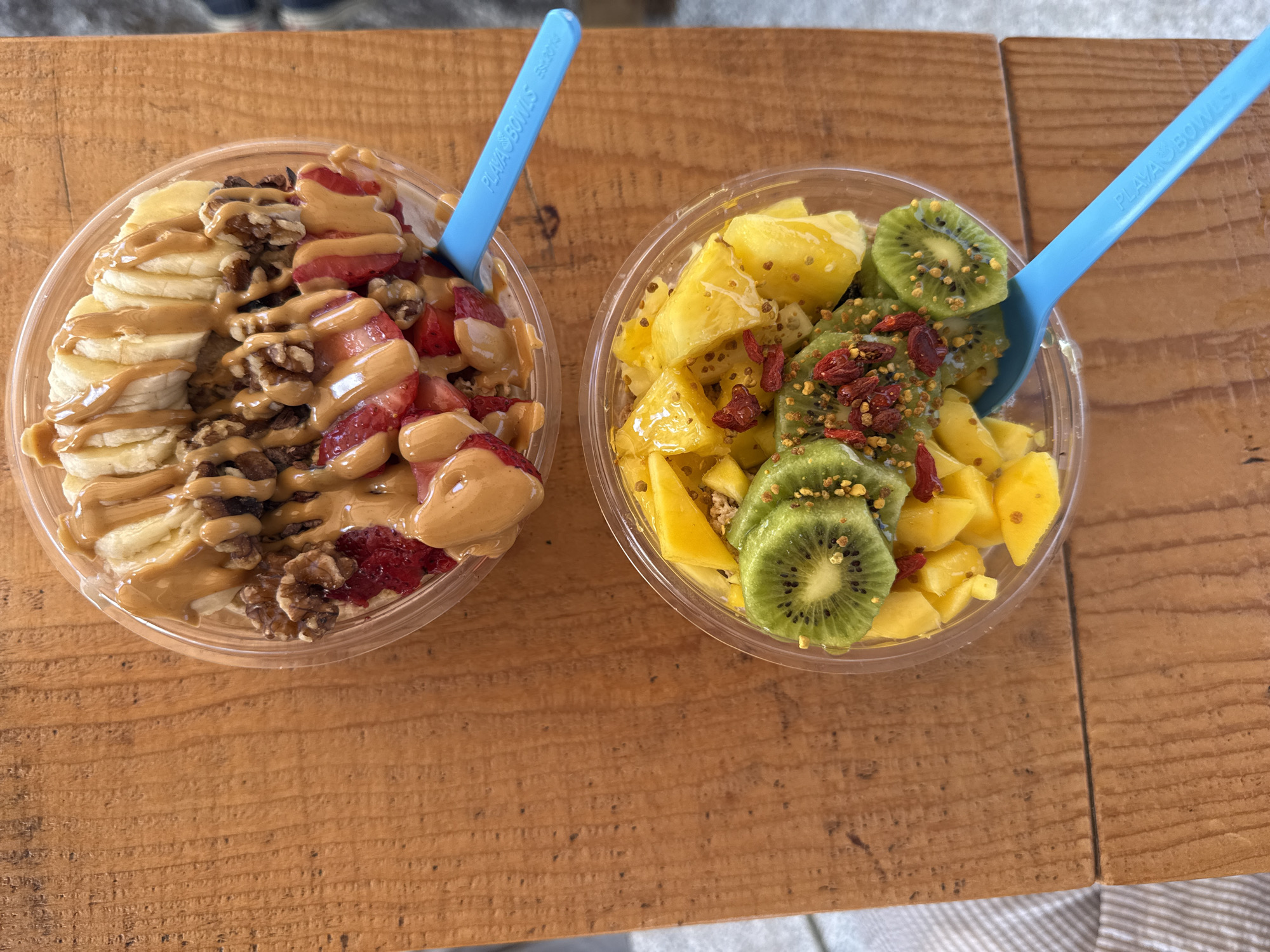 This image shows two fruit bowls on a wooden surface, likely a table. The bowls appear to be made of clear plastic.
The bowl on the left contains sliced bananas, strawberries, and walnuts, all drizzled with a beige-colored sauce (likely peanut butter). A blue plastic spoon with the words "PLAYA BOWLS" is inserted into the bowl.
The bowl on the right contains diced pineapple and kiwi, also with a blue "PLAYA BOWLS" spoon. The fruit is garnished with small, reddish-brown pieces (likely goji berries) and tiny yellow granules.
The scene is likely indoors, judging by the lighting. The wooden surface is light brown with a visible grain. A patterned material is visible underneath the table.
The image appears to have been taken in Saint Augustine, Florida because Playa Bowls has a location there.