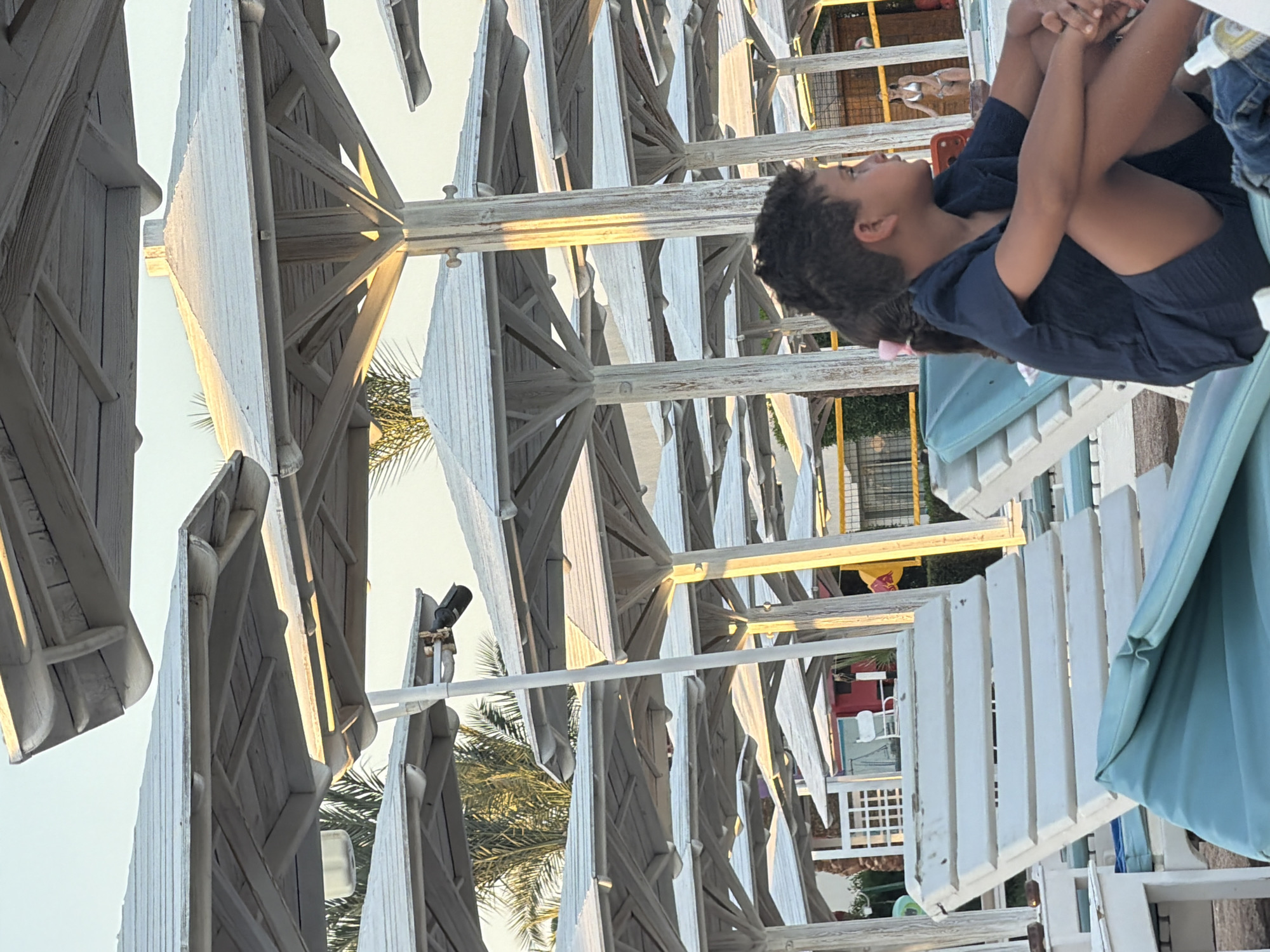 This media file captures a scene at what appears to be a resort or beach area in Sharm El Sheikh, Egypt. The primary subjects are two young children, a boy and a girl, positioned in the foreground. The boy, with curly dark hair, is seated with his knees drawn up to his chest, looking upwards and to his left with a contemplative expression. He is wearing a dark blue t-shirt and shorts. A girl, partially visible, is sitting behind him, also looking upwards.
The background is dominated by rows of white wooden structures that resemble beach cabanas or gazebos, with white fabric canopies. Sunlight filters through these structures, casting a warm glow and creating dappled light. Palm fronds are visible interspersed among the structures, suggesting a tropical or semi-tropical environment. In the distance, some structures with red and yellow accents can be seen, possibly part of the resort's facilities.
The image is oriented vertically, with the children appearing on the right side and the cabanas extending from the bottom to the top. The time of day appears to be late afternoon or early evening, given the warm, low-angled sunlight. The sky is bright and clear, indicating fair weather. There is no visible text within the frame. The overall mood is one of relaxation and quiet observation, possibly during a family vacation.