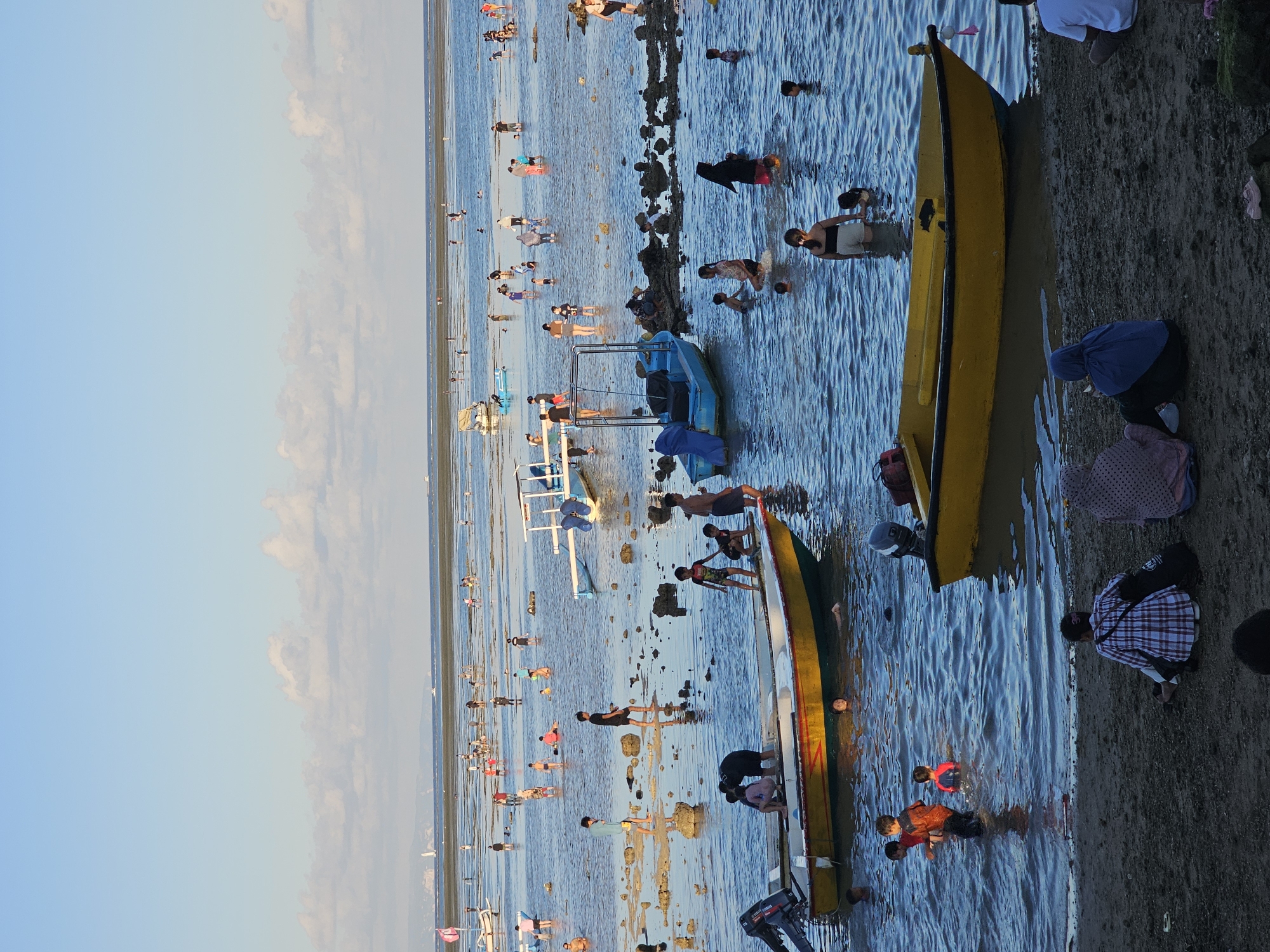 This outdoor shot depicts a crowded beach scene at Sanur Beach in Denpasar, Indonesia. The sky is partially cloudy. Numerous people are wading in the shallow water at low tide, many near several small boats. Two yellow boats lie close to the shore. In the immediate foreground, several individuals stand near the boats, while others are knee-deep in the water. The beach extends into the distance, populated with numerous people enjoying the water.