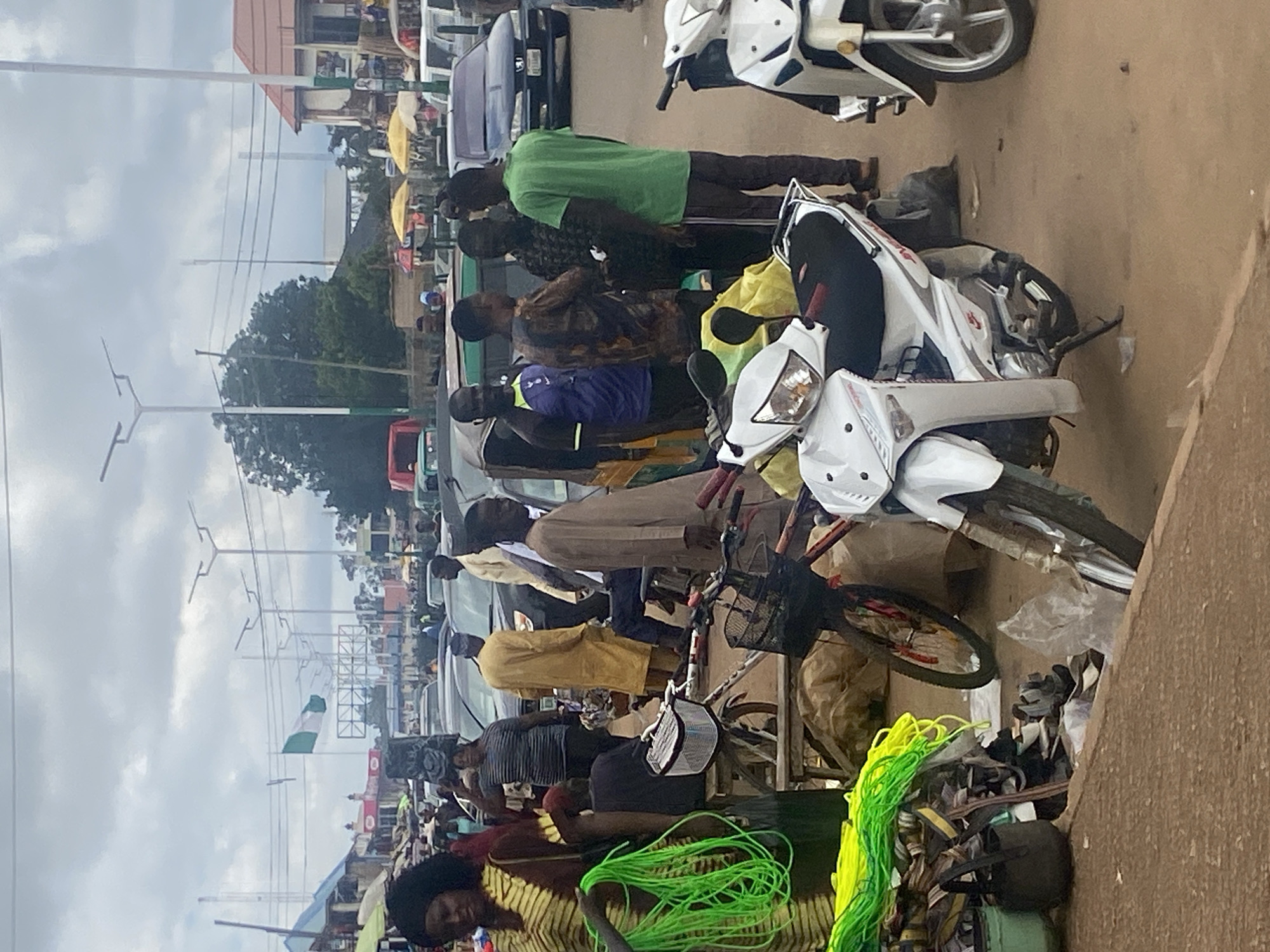 This image depicts a street scene in Bukuru, Nigeria, likely a market area. A group of individuals are gathered near parked motorcycles and a bicycle. In the foreground, a woman in a patterned dress holds a bundle of bright green ropes. To her right, a white motorcycle is prominently displayed. Behind the woman, a bicycle with a basket is visible. Several men are interacting in small groups, some engaged in conversation. One man in a green t-shirt stands facing others, while another in a yellow robe walks away. The environment features numerous parked vehicles, including cars and motorcycles, suggesting a busy commercial or transportation hub. Streetlights and some buildings are visible in the background, under a cloudy sky. A Nigerian flag is also visible in the distance, flying from a pole. The ground appears to be a paved or dirt road surface.
