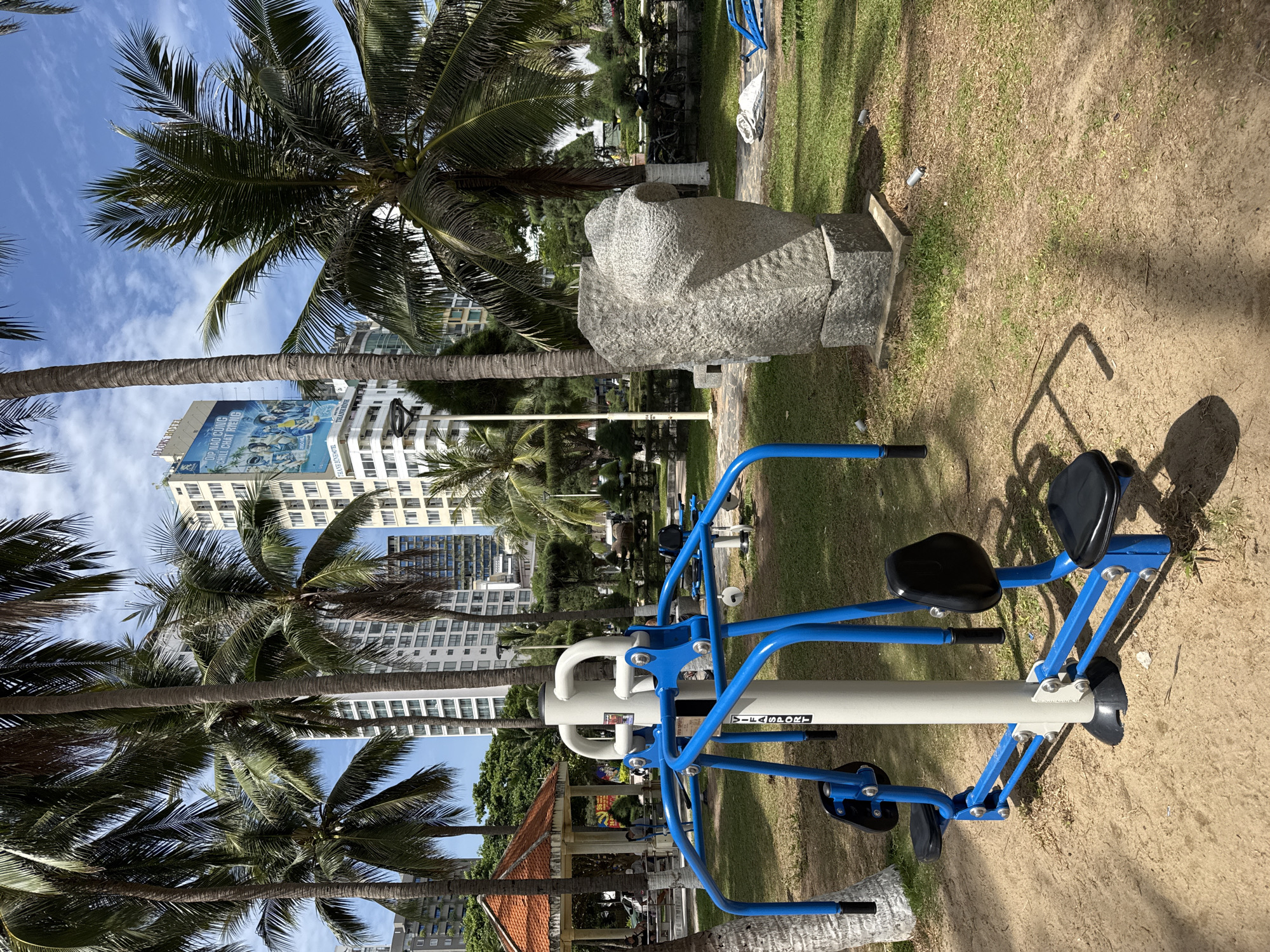 The image depicts an outdoor fitness area in Nha Trang, Vietnam, characterized by a clear blue sky with scattered clouds and lush green vegetation. In the foreground, a blue and white outdoor exercise machine, likely for chest presses or similar movements, is prominent. The machine has black padded seats and handles. Behind the fitness equipment, a large, gray, rough-hewn stone sculpture resembling a stylized human head stands on a concrete base.
Further in the background, several tall palm trees sway gently, their fronds creating dappled shadows on the ground. Beyond the trees, modern high-rise buildings, typical of a coastal city, rise against the sky. One building, identified as "GLORY HOTEL" by signage, features a large advertisement on its facade. A small, orange-roofed pavilion is also visible, suggesting a public park or recreational space. The ground is a mix of sand and short, dry grass, indicating a warm, possibly tropical climate. Several individuals can be seen in the mid-ground, seated and conversing under the pavilion, and some are walking or standing near other fitness equipment. The overall scene suggests a well-maintained public space designed for leisure and exercise.
