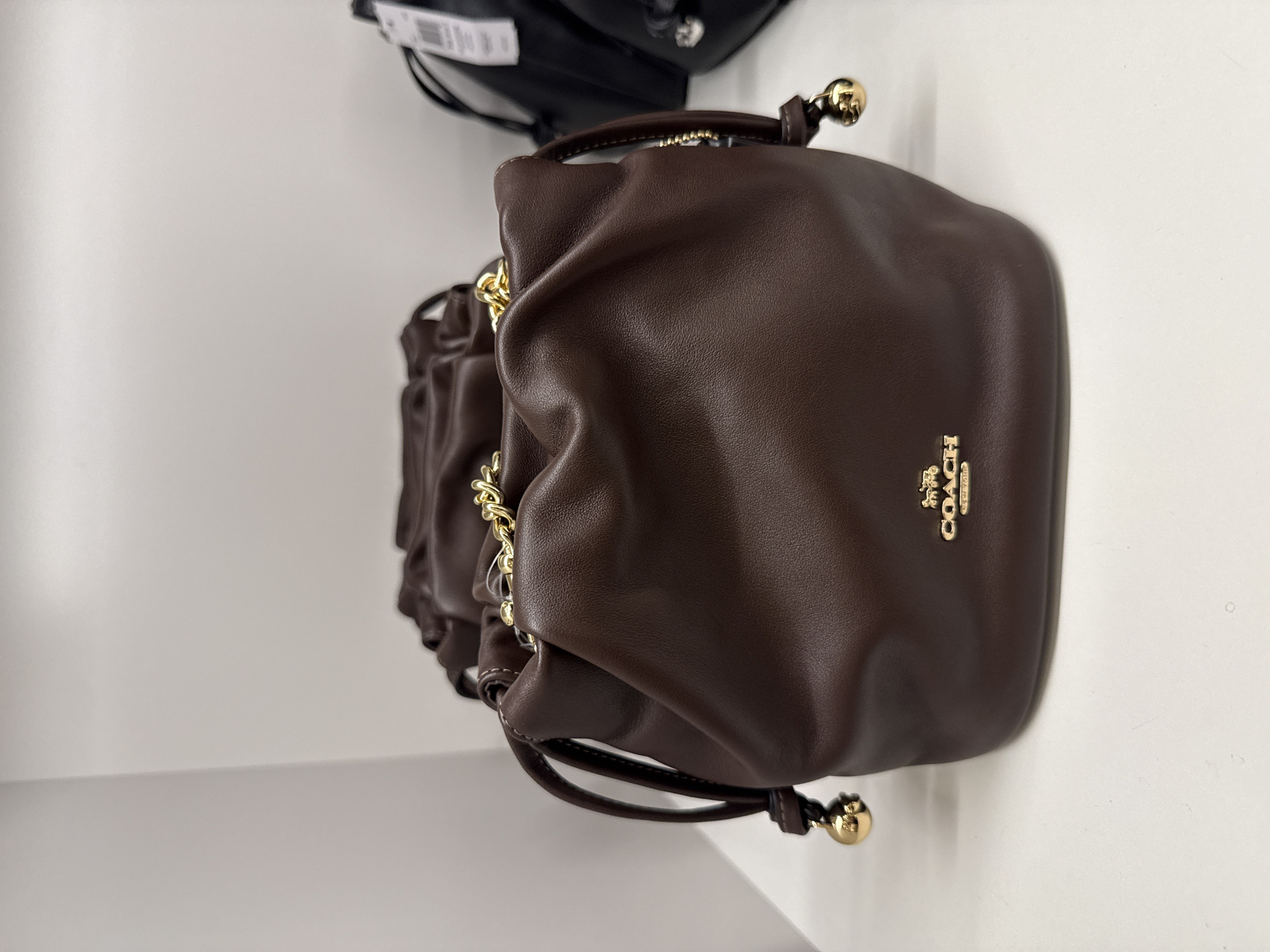 The image shows a brown Coach drawstring bag with the Coach logo on its front. The bag is sitting on a white surface, likely a shelf or display case. The picture was probably taken indoors, and there is no indication that it was taken in Sunrise, United States of America.