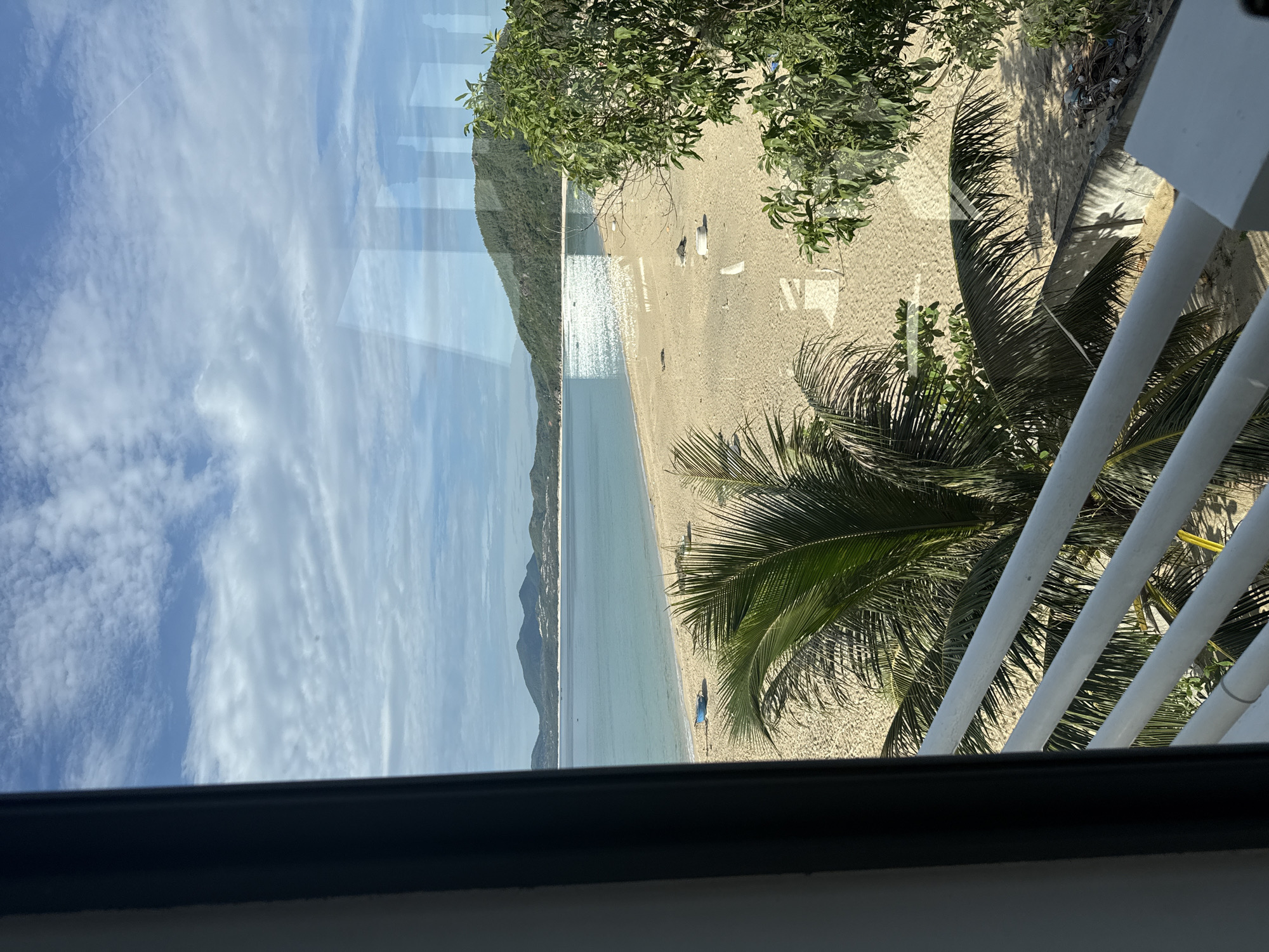 A serene beach scene unfolds beyond a window, offering a picturesque view of the ocean and a sandy shoreline. In the foreground, a white railing and lush green palm fronds partially obscure the view, hinting at a balcony or veranda. The sand is a light golden color, with a few scattered objects, including what appears to be a blue boat resting near the water's edge. Beyond the beach, the turquoise water stretches out towards a distant coastline marked by rolling green hills and mountains under a bright blue sky dotted with white clouds. Sunlight glints off the water, creating a shimmering effect. The overall impression is one of tranquility and natural beauty.