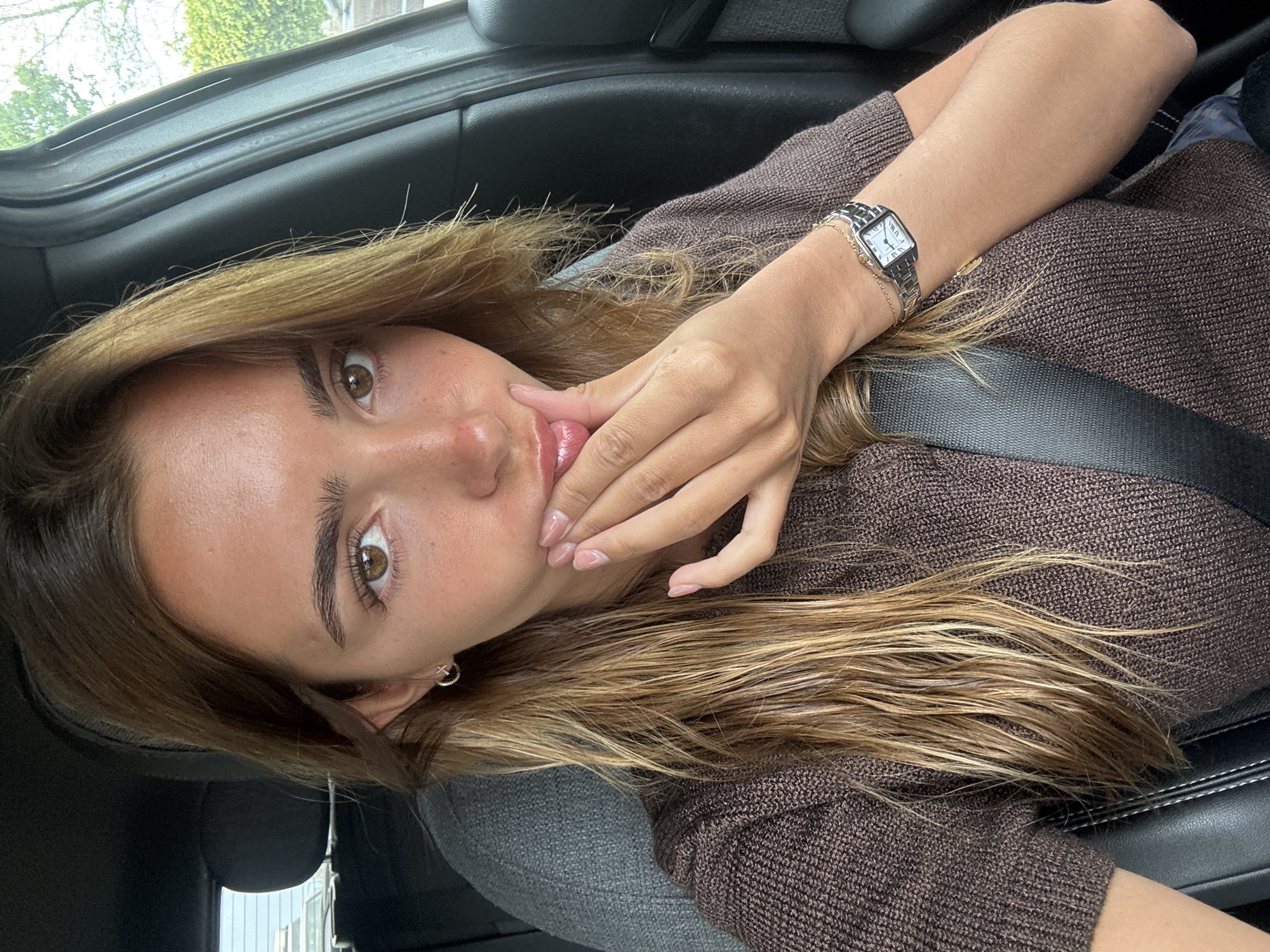 A young woman is inside a car. She wears a brown knitted sweater. A watch is on her left wrist. She has fair skin, brown hair with blond highlights, and light brown eyes. She has a seatbelt across her torso. She is holding her hand up to her mouth. The car's interior is grey and black. The image provides no information to suggest a location of the city of Porto, Portugal.