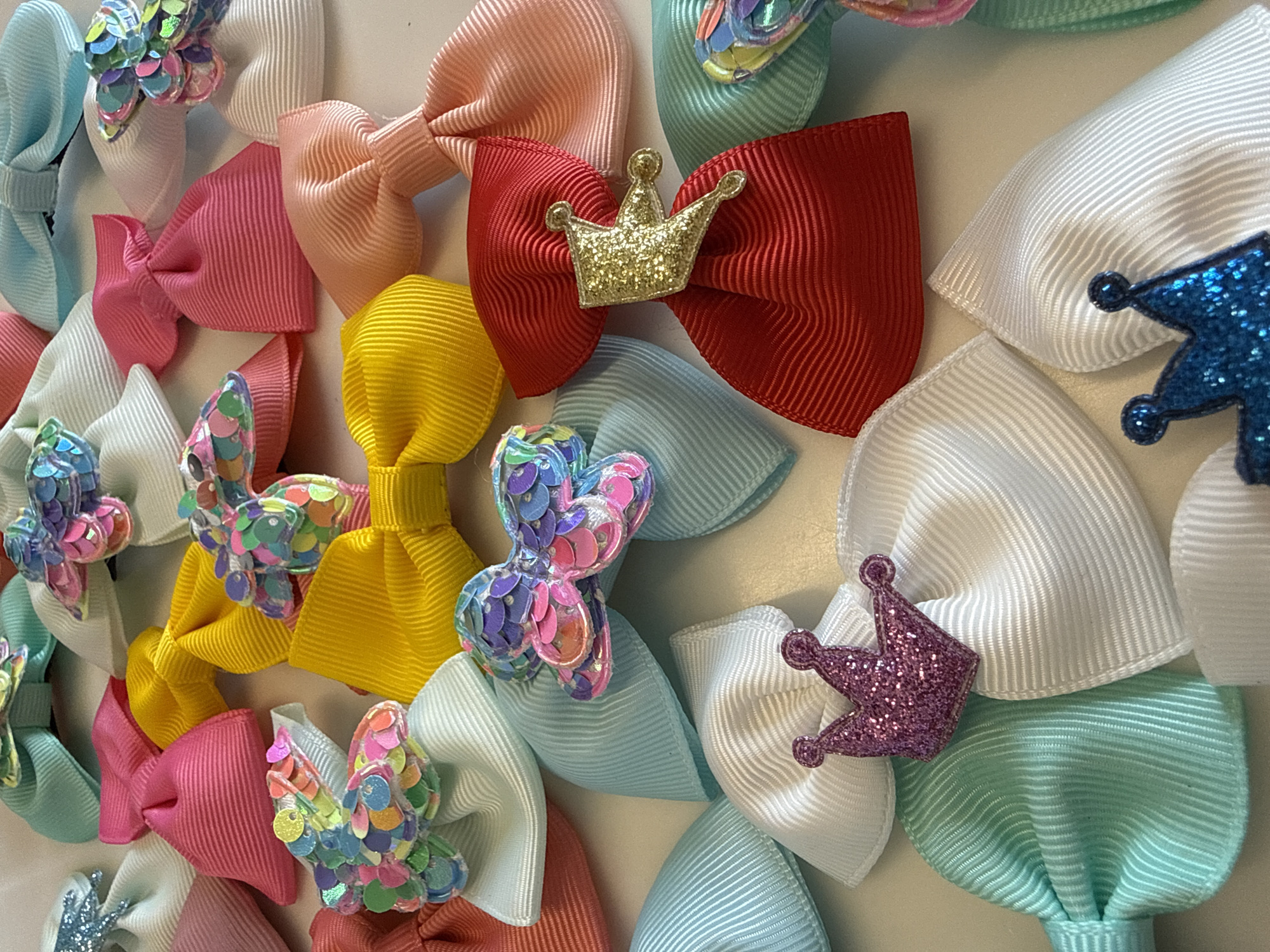 The image shows a collection of colorful hair bows arranged on a flat surface. The bows are made of ribbon and adorned with decorative elements such as glittery butterflies and crowns. The bows vary in color, including pink, red, yellow, blue, and white. The arrangement appears to be random. The setting is unclear and there is no visible location of Votkinsk, Russia, evident in the picture.