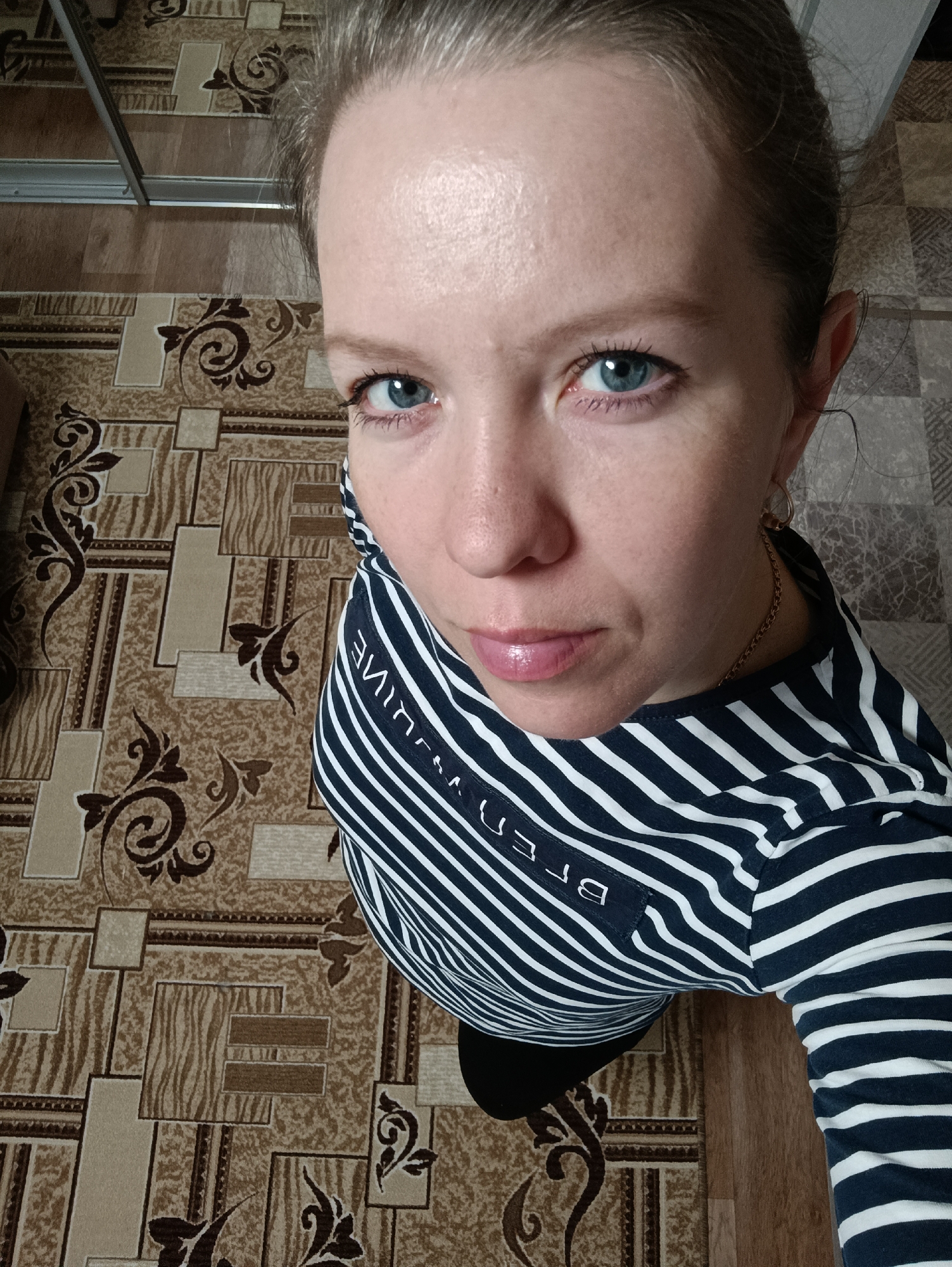 A close-up, high-angle portrait features a woman indoors in Chaikovsky, Russia, looking directly at the camera. Her bright blue eyes are the focal point, complemented by a neutral expression with slightly parted lips. Her hair is pulled back from her face. She is wearing a navy blue and white striped long-sleeved shirt, with a thin gold chain necklace and a small hoop earring visible on her right side. Underneath the striped top, a black garment is partially visible. On her left sleeve, partially visible, upside-down, and mirrored text resembling "ВЛИН E" or similar letters is present, likely a brand or design element. The background is a patterned floor, possibly linoleum or carpet, featuring an intricate design of brown and beige geometric shapes and ornate floral motifs. A metallic rail or part of furniture is subtly visible in the top left corner.