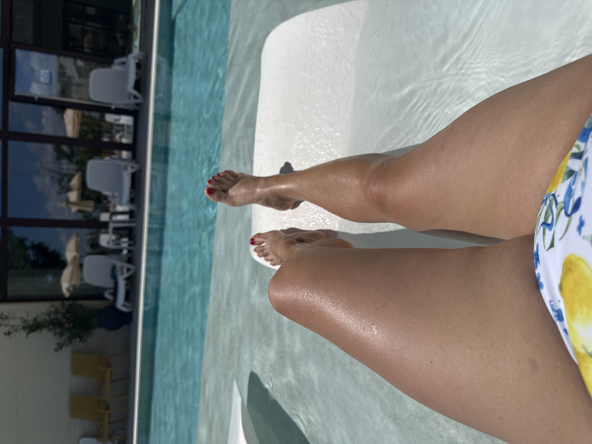 A first-person perspective shot shows a person's legs and feet relaxing in shallow water at the edge of a swimming pool. The person's legs are tan and appear to have been recently in the sun, with visible water droplets on the skin of their left leg. Their toenails are painted red. They are wearing a bikini bottom with a lemon and floral pattern. The pool water is clear and blue, with gentle ripples on the surface. In the background, several white lounge chairs are arranged around the pool, and behind them are large glass windows reflecting the sky and trees. The scene suggests a leisurely day by a pool at a hotel or resort. The location is unknown.