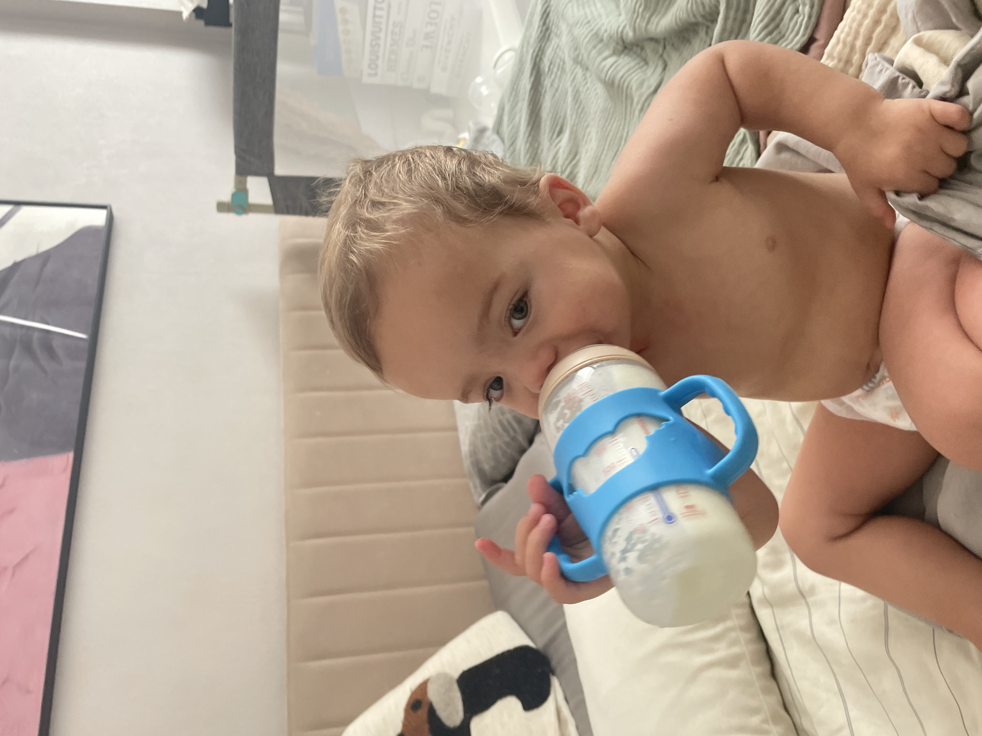 A toddler, with blond hair and blue eyes, is sitting on a bed, holding and drinking from a baby bottle with a blue handle. The toddler is wearing a diaper. The bed is covered with a striped sheet and a green textured blanket. A headboard is visible behind the toddler, with a decorative pillow on the left side. In the background, a crib with a grey railing is seen, and on a shelf behind it are several books or decorative boxes with brand names like "LOUIS VUITTON" and "HERMES." The scene appears to be indoors, likely in a bedroom. There are no visible indicators of the city of Almaty, Kazakhstan.