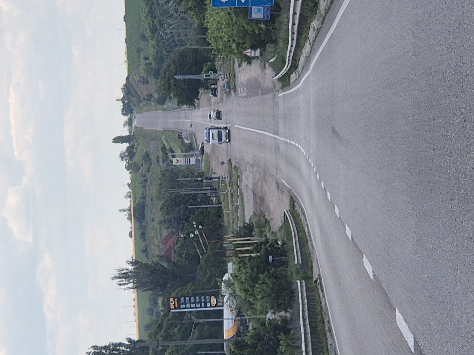 The image shows a road leading uphill in Pidhaitsi, Ukraine. On the left side of the road, there is a gas station with a price display sign. Further up the road, there are several vehicles, including a truck and cars. The landscape on either side of the road is hilly and covered with greenery and some trees. The sky is partly cloudy.