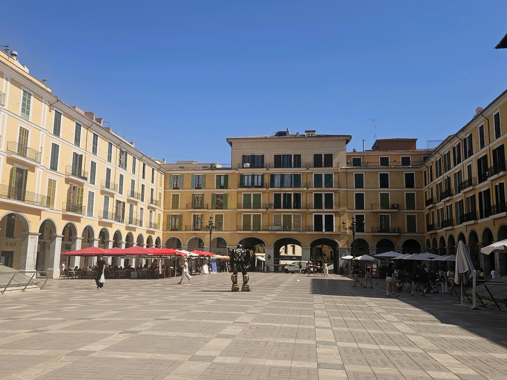 The image depicts a sunny day in the Plaça Major of Palma, Spain. The square is surrounded by historic buildings with yellow facades, arched ground floors, and numerous windows with dark shutters. In the foreground, the tiled pavement is laid out in a geometric pattern.
In the center of the square, a person in a robot costume, resembling Bumblebee from Transformers, stands stationary. Several people are scattered across the plaza, some walking and others seated at tables under red umbrellas, suggesting outdoor dining. A woman in a white dress walks across the square, and another person, dressed in black, carries a white bag. Other individuals are visible in the background, near cafes and along the building arcades. The sky is clear and blue, indicating fair weather. The scene conveys a lively, public atmosphere in a European city square.