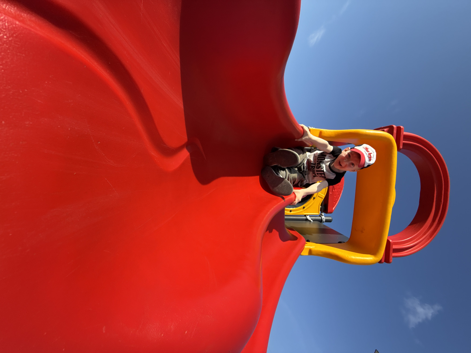 A young boy wearing a red and white baseball cap and a black t-shirt with the words "MONSTER TRUCK" is sitting on a red plastic slide at a playground under a clear blue sky. He is looking towards the camera with a wide smile. The slide is part of a larger playground structure that includes yellow and red plastic elements. The boy's legs are bent, and his feet are resting on the slide. The image is taken from a low angle, emphasizing the curve of the slide. The location in Vereshchagino, Russia, is not discernible from the image itself.