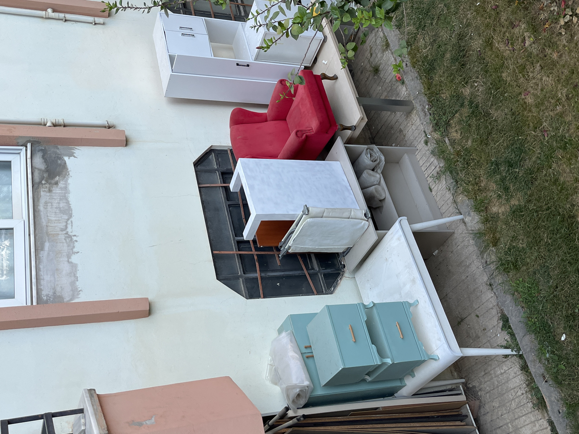 In Alanya, Turkey, a collection of furniture is staged against a building's pale yellow exterior wall, near a partially overgrown grassy area. The scene appears to be an outdoor storage or staging area.
Dominating the center-right of the frame is a vibrant red armchair with plush upholstery and ornate wooden legs. To its left, a white, rectangular cabinet with two doors and open shelving stands. Adjacent to this is a tall, white, narrow cabinet with two doors and visible shelving inside.
In the foreground and mid-ground, several other furniture pieces are arranged. A light-colored, rectangular table with four slender white legs supports a stack of two teal-colored drawers, each with a wooden knob. A white sheet is draped over the top drawer. To the right of these drawers is a white, open-shelf unit. Further right, a light brown surface appears to be a shelf or table.
Behind the red armchair, a dark, recessed opening in the wall is visible, possibly a window or ventilation shaft, with a metal grate. The building features architectural elements such as vertical pink trim around windows and conduits running down the wall.
The ground is a mix of concrete paving and natural grass, with some fallen leaves visible. The overall impression is one of discarded or temporarily stored household items.