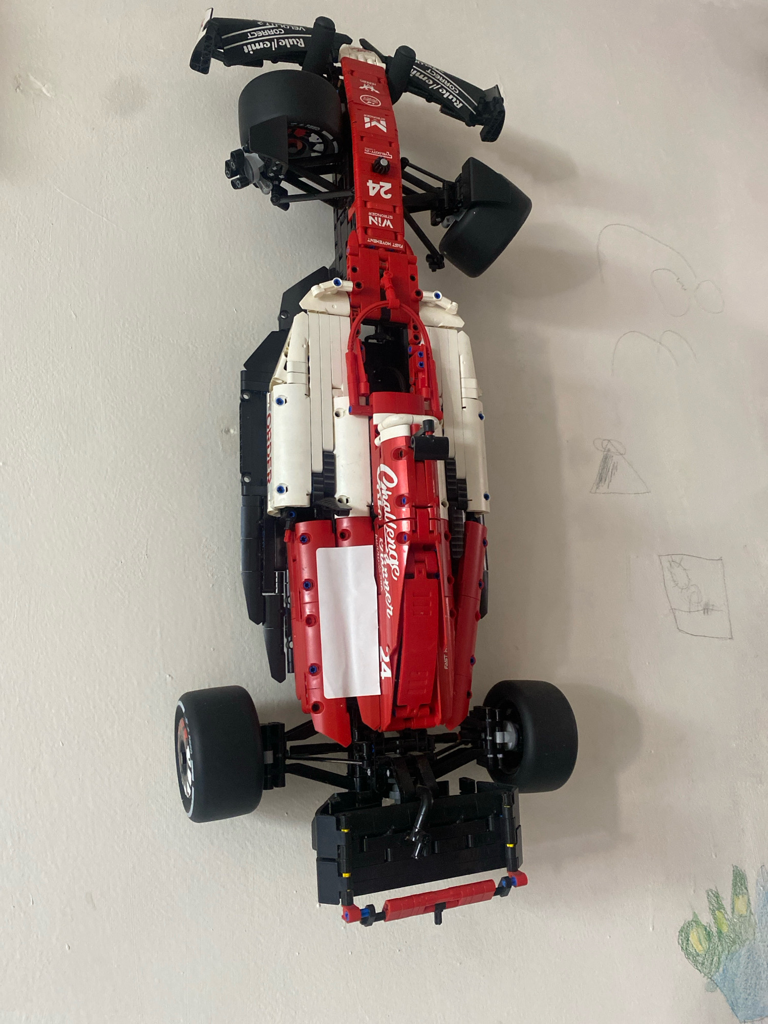 A red and white Lego Technic Formula 1 race car is mounted vertically on a white wall. The car is viewed from above, with its front pointing upwards. The car has black tires with white sidewalls, a red chassis, and white sidepods. There are various decals on the car, including the number 24, and what appear to be sponsor logos. To the right of the car, there are faint child-like drawings on the wall, including a few curved lines, a triangular shape with shading, and a rectangular drawing with some scribbled elements. The wall appears textured and slightly uneven. The lighting suggests an indoor environment, likely a room. There are no people visible in the image. There is no information within the image that would indicate the location is Bandar Lampung, Indonesia.