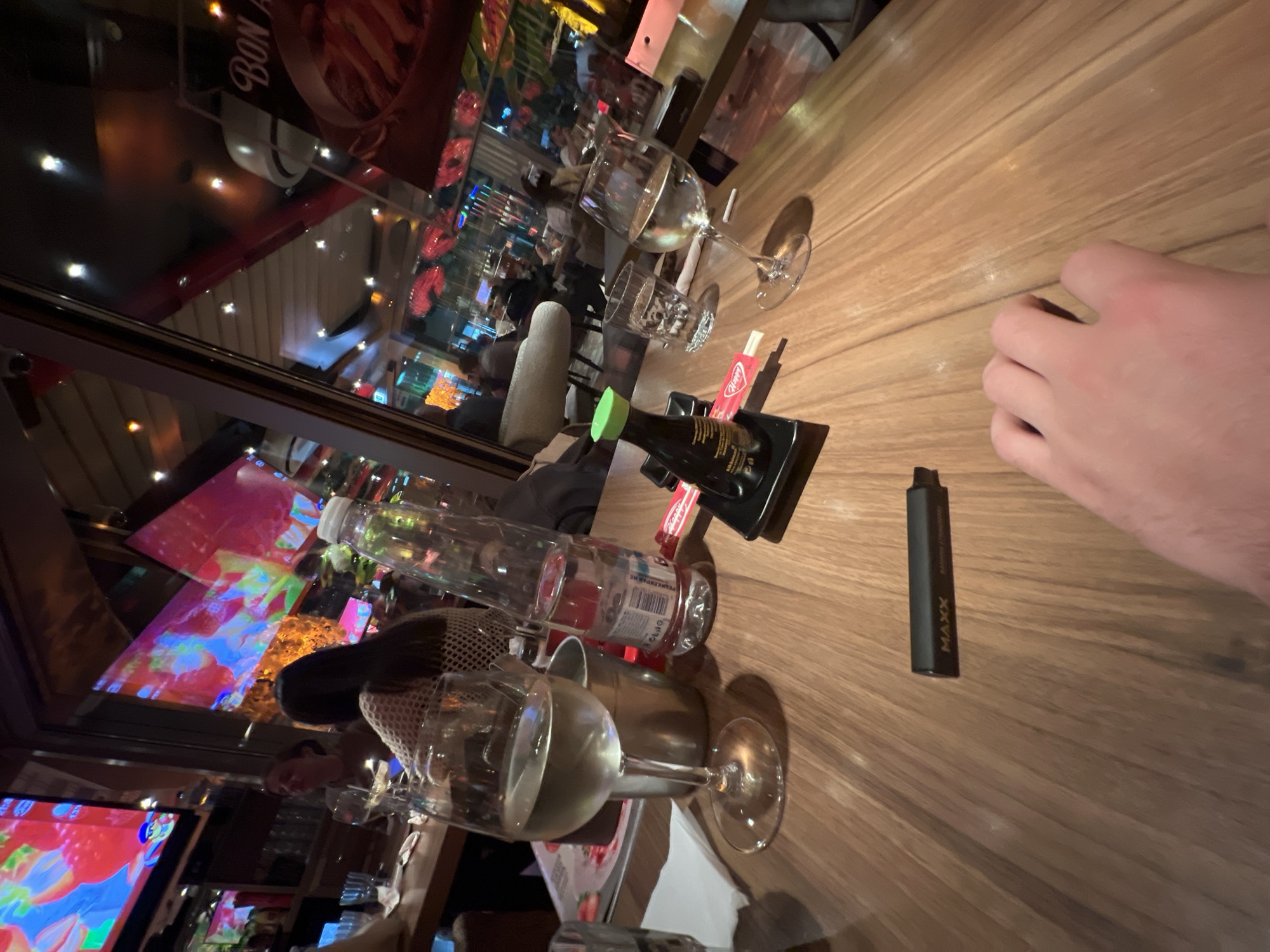 The scene is a table at a restaurant in Sofia, Bulgaria. The table is light brown, and there are several objects on it. From left to right, there is a wine glass, a water bottle, a soy sauce bottle, and a black vape pen. A person's hand is also visible. The restaurant has a window and interior lighting is visible. There are reflections visible in the window. Other people and objects are blurred and out of focus.