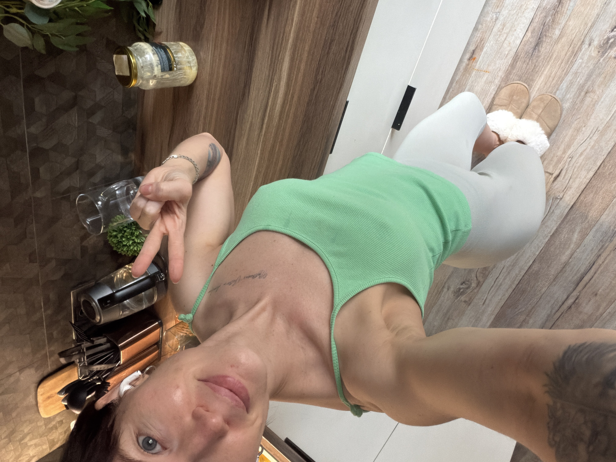 A rotated, tilted, close-up selfie of a woman wearing a light green ribbed tank top and light gray leggings or pants. She is wearing tan slippers with white fur. Her left hand is raised with fingers in a peace sign. Her left arm has a visible tattoo. She has short dark hair and blue eyes. The location appears to be a kitchen with wood-look cabinets and countertops. The background contains a jar of pickled goods, a vase with greenery, a knife block with utensils, and a small succulent. The environment suggests a domestic interior. There is no information available to verify the location in the city of Perm, Russia.