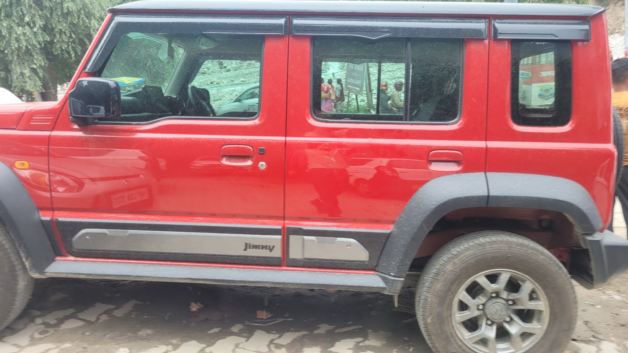 This image shows the side of a bright red, four-door vehicle, likely a Suzuki Jimny, parked on a street. The car has a black roof and black fender flares. The doors and windows are visible, with reflections of trees, people, and a sign in the windows. A small orange indicator light is on the front fender. The words "Jimny" are visible on a silver accent panel on the lower part of the door. The ground appears wet, suggesting recent rain or a wet surface, and the lighting indicates it is daytime. The background shows more greenery and some indistinct structures. The overall scene is likely a street in Vashisht, India, given the context.