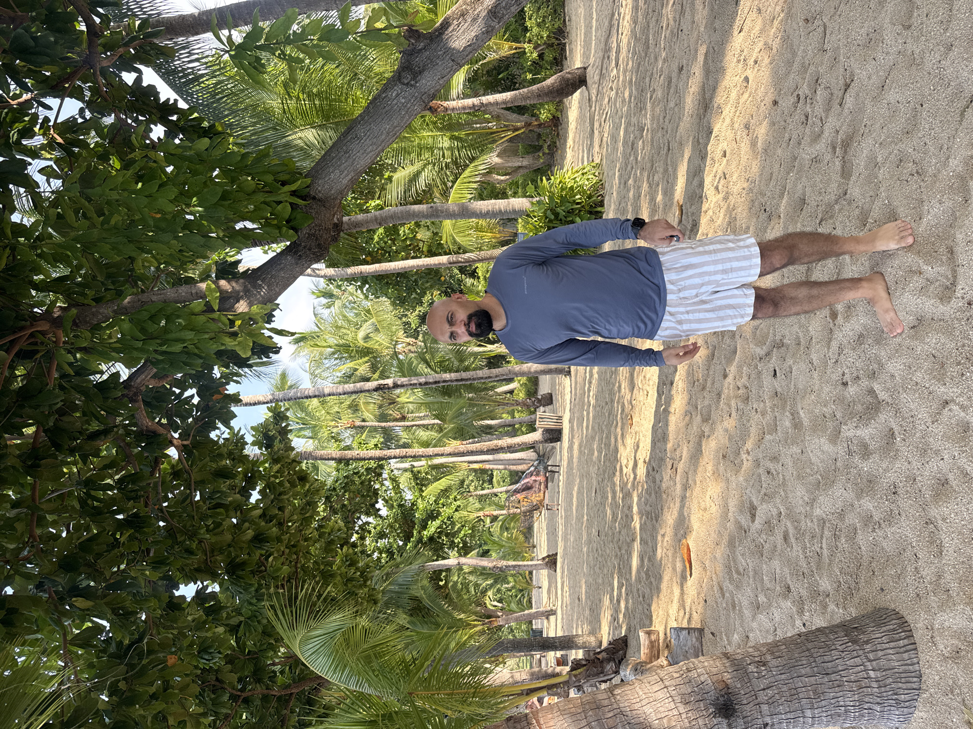 A bald, bearded man with dark skin stands on a sandy beach in front of a line of palm trees. He is wearing a dark blue long-sleeved shirt and white and tan striped shorts. The man is facing to his left, with his head turned further to the left, looking off into the distance. His left arm is slightly bent, and his right arm hangs loosely at his side. There is a hammock visible in the background, strung between two palm trees. The sand is light brown and appears to be dry, with some scattered debris and fallen leaves. The sun is casting long shadows across the sand, indicating it is either early morning or late afternoon. The overall atmosphere is tropical and relaxed. This location appears to be in Ko Samui District, Thailand, given the abundance of palm trees and the sandy beach environment.