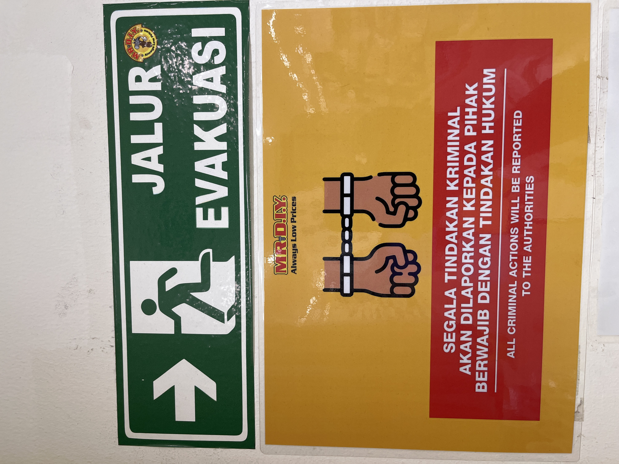 The image shows two signs mounted on a wall. The upper sign is green and features a white arrow pointing to the right next to a white silhouette of a person running. Above the running person, the words "JALUR EVAKUASI" are printed in white, and to the right of "JALUR", a circular logo with the letters "MR.D.I.Y." is visible. The lower sign is predominantly yellow with a red rectangular section at the bottom. The yellow area contains an illustration of two hands shackled together. The red section contains text in white. The top line reads "SEGALA TINDAKAN KRIMINAL", followed by "AKAN DILAPORKAN KEPADA PIHAK" on the next line, and "BERWAJIB DENGAN TINDAKAN HUKUM" on the line below that. Beneath this, in smaller white text, are the English words "ALL CRIMINAL ACTIONS WILL BE REPORTED TO THE AUTHORITIES". The wall appears to be a light color, possibly painted. The signs are presented in a straightforward manner, suggesting they are informational notices within a commercial or public space. The presence of the MR.D.I.Y. logo indicates a potential location within a MR. D.I.Y. store. The text on the signs is a warning about reporting criminal actions and an evacuation route, typical of safety and security signage. There is no specific indication of the city of Tangerang, Indonesia, within the image itself, but MR. D.I.Y. is a well-known chain of home improvement stores present in Indonesia, including Tangerang.