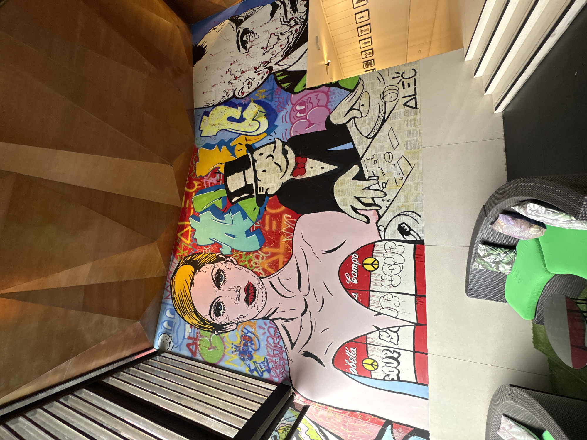 The image shows an interior space with a painted mural on the floor. The mural features a pop art style depiction of various figures including Andy Warhol and Monopoly's Mr. Monopoly, amidst abstract designs and the Campbell Soup logo. Adjacent to the mural is a wooden staircase and metallic seating. The setting is likely a design-conscious space or interior, given the presence of the distinct artwork, Baan Bo Phut, Thailand.