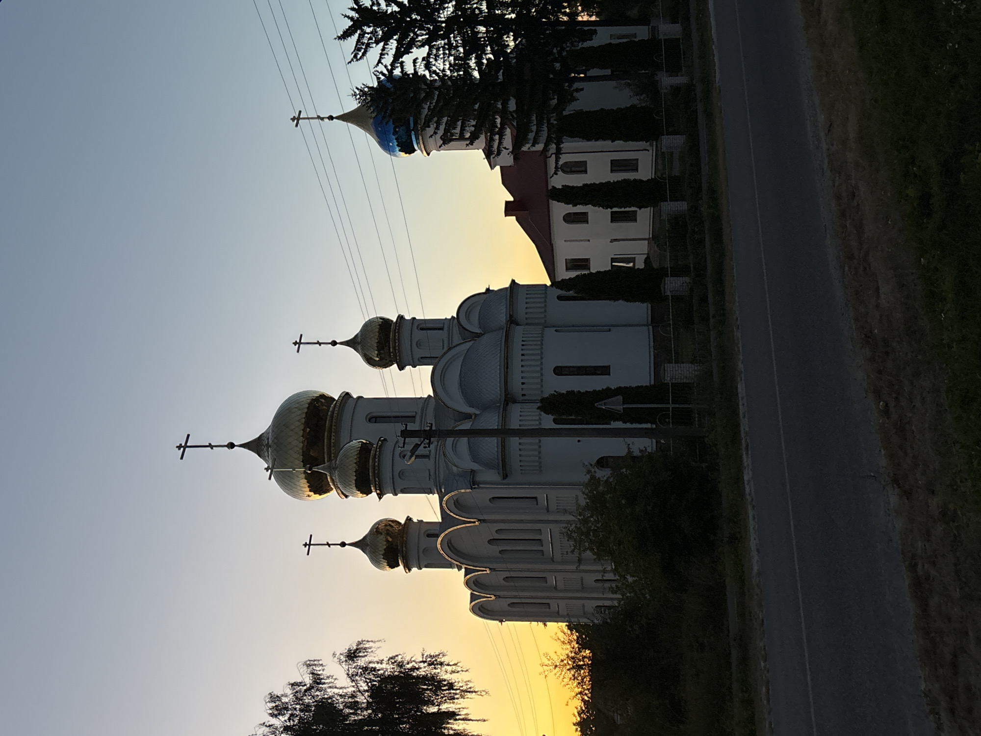 The image shows an Orthodox church with golden domes and crosses in the city of Lanivtsi, Ukraine. The church is white with grey accents and has arched windows. There is a smaller white building with a red roof next to the church, also featuring arched windows. The scene is set against a backdrop of a bright orange and yellow sunset, with a clear blue sky above. Several trees, including a large pine tree, are visible on the right side of the image. A paved road runs in front of the church, with a grassy area and some foliage bordering it. Power lines are visible crossing the sky. The image appears to be taken from across the road, looking towards the church.