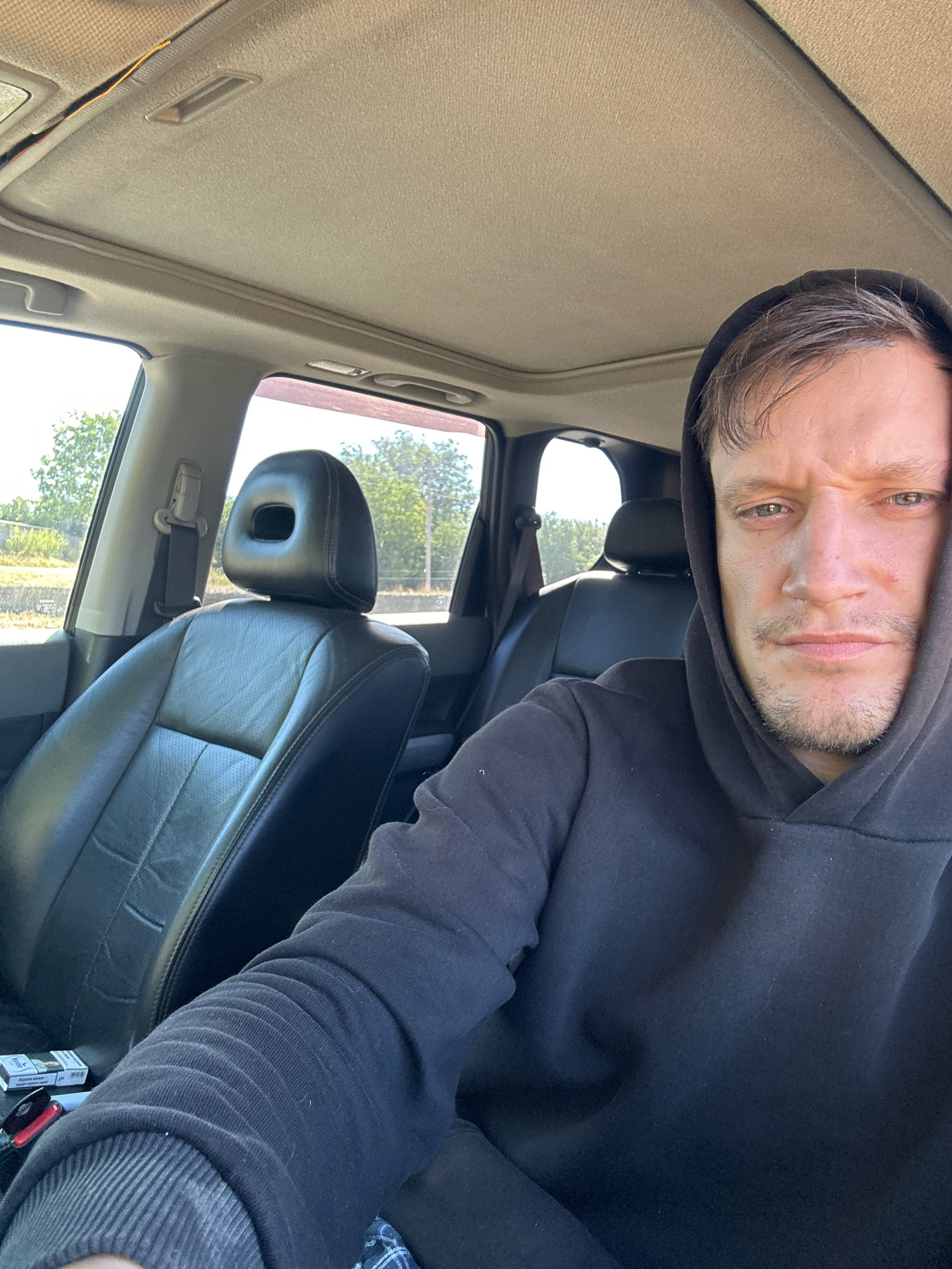 In the city of Makiv, Ukraine, a close-up, waist-up selfie is taken from the perspective of a person sitting in the driver's seat of a car. The car's interior, a tan headliner and black leather seats, is visible. The person, wearing a black hoodie, has their right arm resting on the steering wheel and their left arm is not visible. A pack of cigarettes and a lighter are on the seat beside them. The person's face is directed towards the camera, with an unexpressive look. The background shows trees and greenery, suggesting an outdoor location.