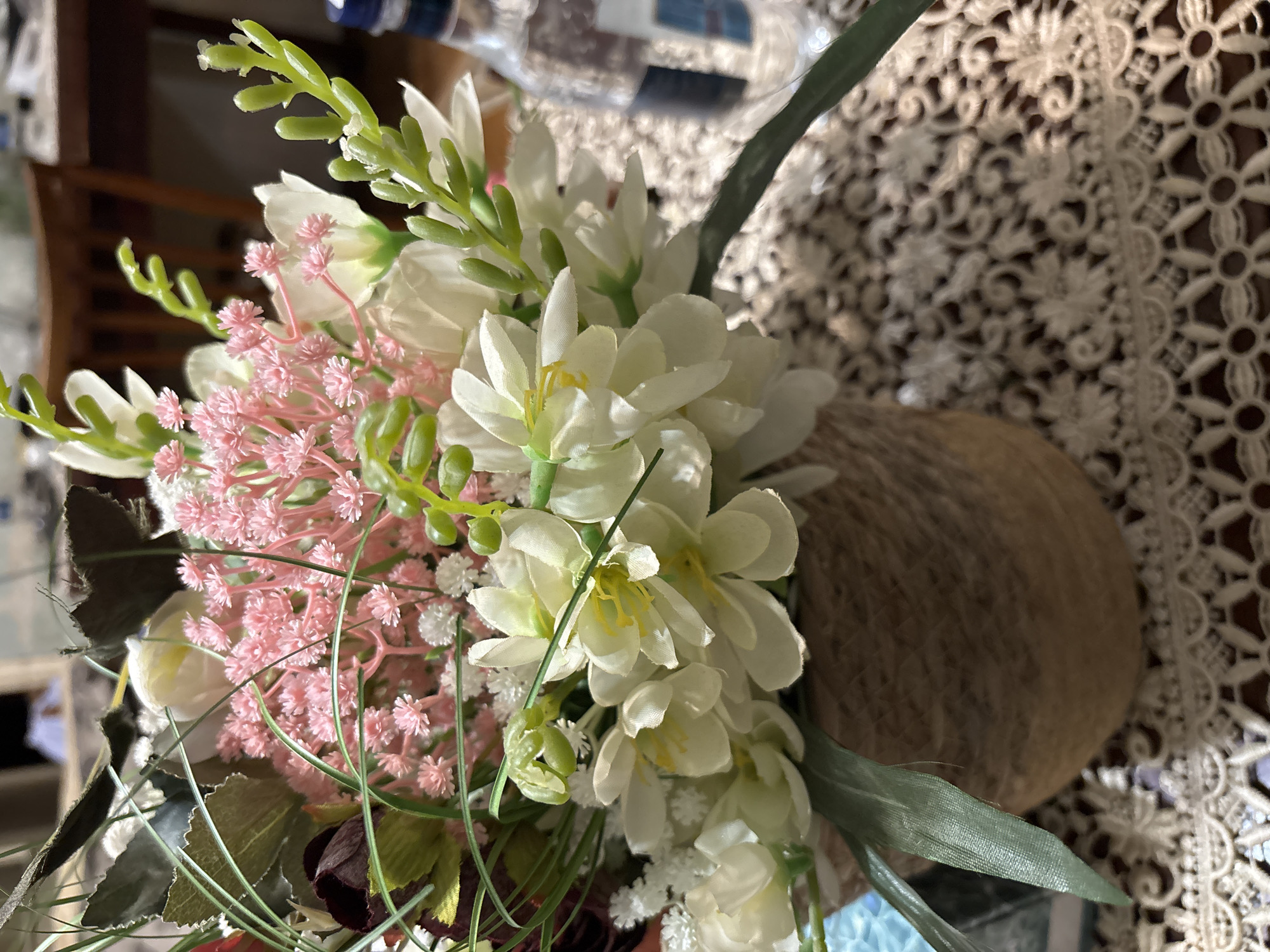 The image displays an artificial floral arrangement, likely placed indoors. The main subject is a bouquet consisting of white and pink flowers, interspersed with green foliage. The white blossoms are prominent, featuring visible yellow stamens and petals resembling freesia or similar delicate flowers. Clusters of small, light pink flowers add a soft contrast. Various green stems and leaves provide a natural backdrop to the blooms.
The flowers are held in a round, brown container, which appears to be wrapped with twine or a similar textured material, giving it a rustic aesthetic. This container rests on a surface partially covered by a white lace tablecloth. The tablecloth has an intricate, openwork floral pattern. The background is softly blurred, showing hints of dark furniture or interior elements. No specific activity or event is taking place, nor are there any people present. The scene conveys a decorative and tranquil atmosphere, with no visible text. The time of day and weather are not discernible from the indoor setting.