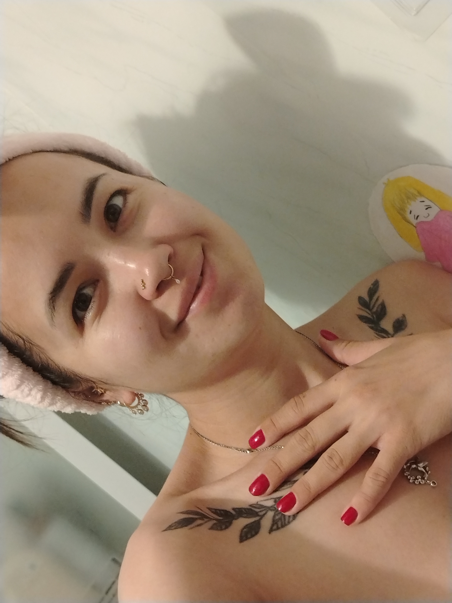 A medium close-up selfie shows a young woman with dark hair pulled back by a pink terrycloth headband. She is looking at the camera with a slight smile and her eyes are partially closed. She has a nose ring and small earrings. A black leaf tattoo is visible on her shoulder. Her nails are painted red, and her hand rests on her chest, covering part of another leaf tattoo. A necklace with a pendant is also visible. In the background, a shadow of a person is cast on a light-colored wall. To the right of the frame, a small circular picture of a person with blonde hair in a pink shirt is attached to the wall. The environment appears to be indoors, possibly a bathroom, given the terrycloth headband and the overall lighting. The image does not provide any information about the city of Noviy, Russia.