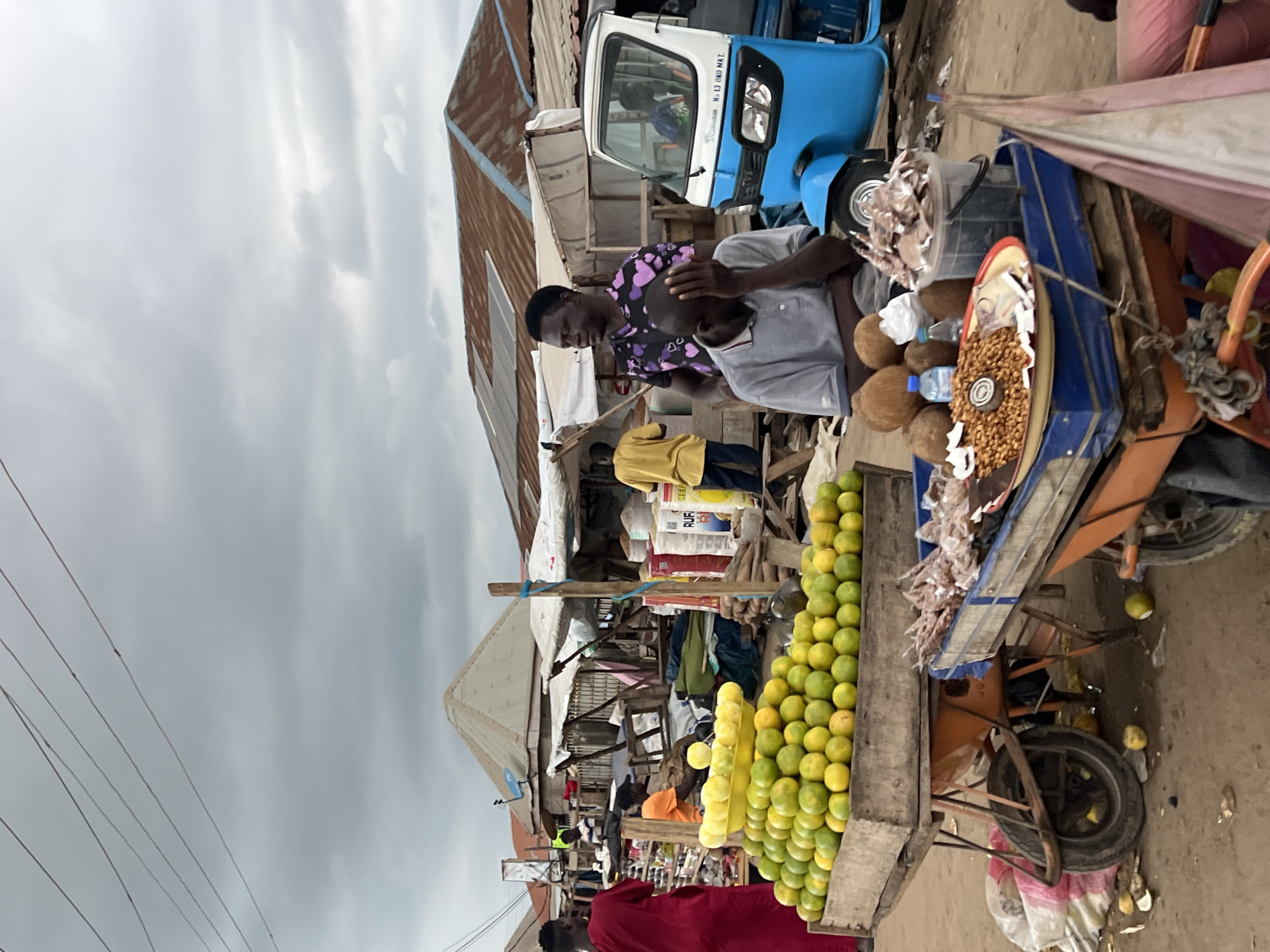 A street market scene unfolds in Oko Amakom, Nigeria, under an overcast sky. In the foreground, a wooden cart overflows with a vibrant display of oranges, neatly stacked in a yellow plastic basin. To its right, another cart holds various goods, including a platter of nuts, coconuts, and a bucket of what appears to be dried fish. A blue and white motorized tricycle, commonly known as an "okada" or "kekeno," is parked nearby.
Several individuals populate the scene. A man in a grey polo shirt sits with his head in his hand, seemingly dejected, next to the tricycle. Another man with a purple shirt adorned with hearts stands behind him, looking towards the left. To the far left, a person in a bright red garment stands facing away from the camera. In the mid-ground, a person in a yellow shirt is visible standing near a stall. The market environment is characterized by makeshift stalls with corrugated metal roofs and tarpaulin coverings, typical of informal markets in the region. Wires are visible strung overhead. The ground is dusty and uneven, with some scattered debris. The overall atmosphere suggests a bustling, albeit somewhat subdued, market day.