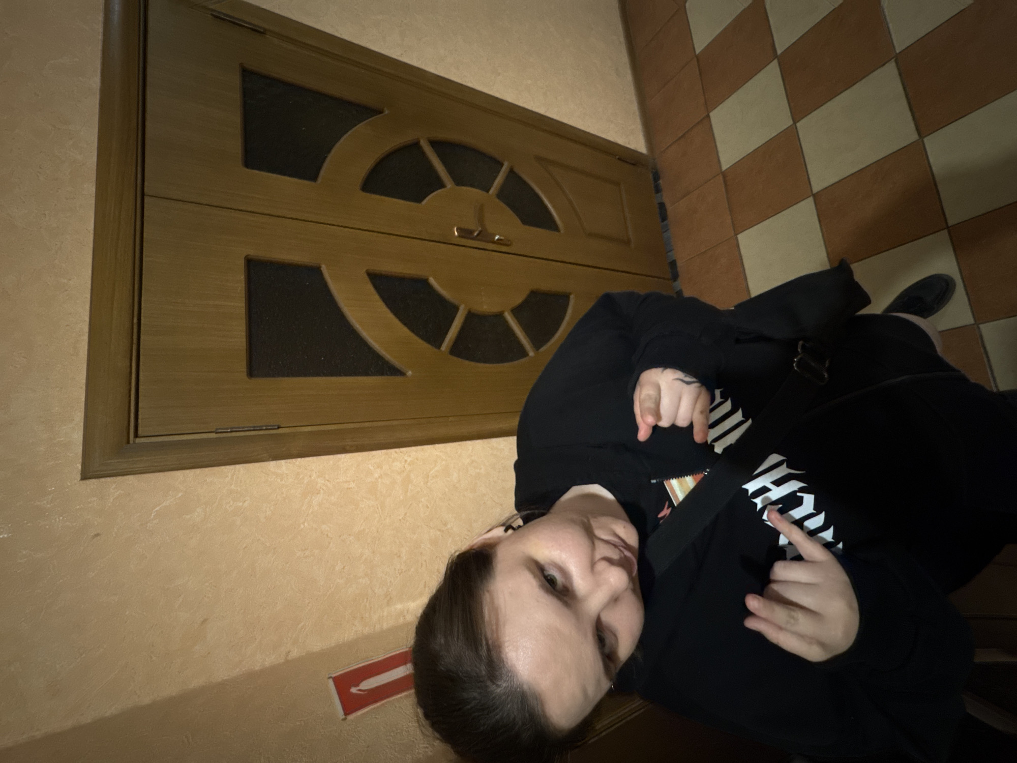 A person is posing for a photo in what appears to be an indoor hallway. The person is wearing a black hoodie and has their left hand raised with two fingers extended, while their right hand is pointing towards the camera. Behind the person, there are double wooden doors with glass inserts and a beige-colored wall. The floor is tiled with a checkerboard pattern of brown and beige squares. A red and white sign is visible on the wall to the left of the doors.
