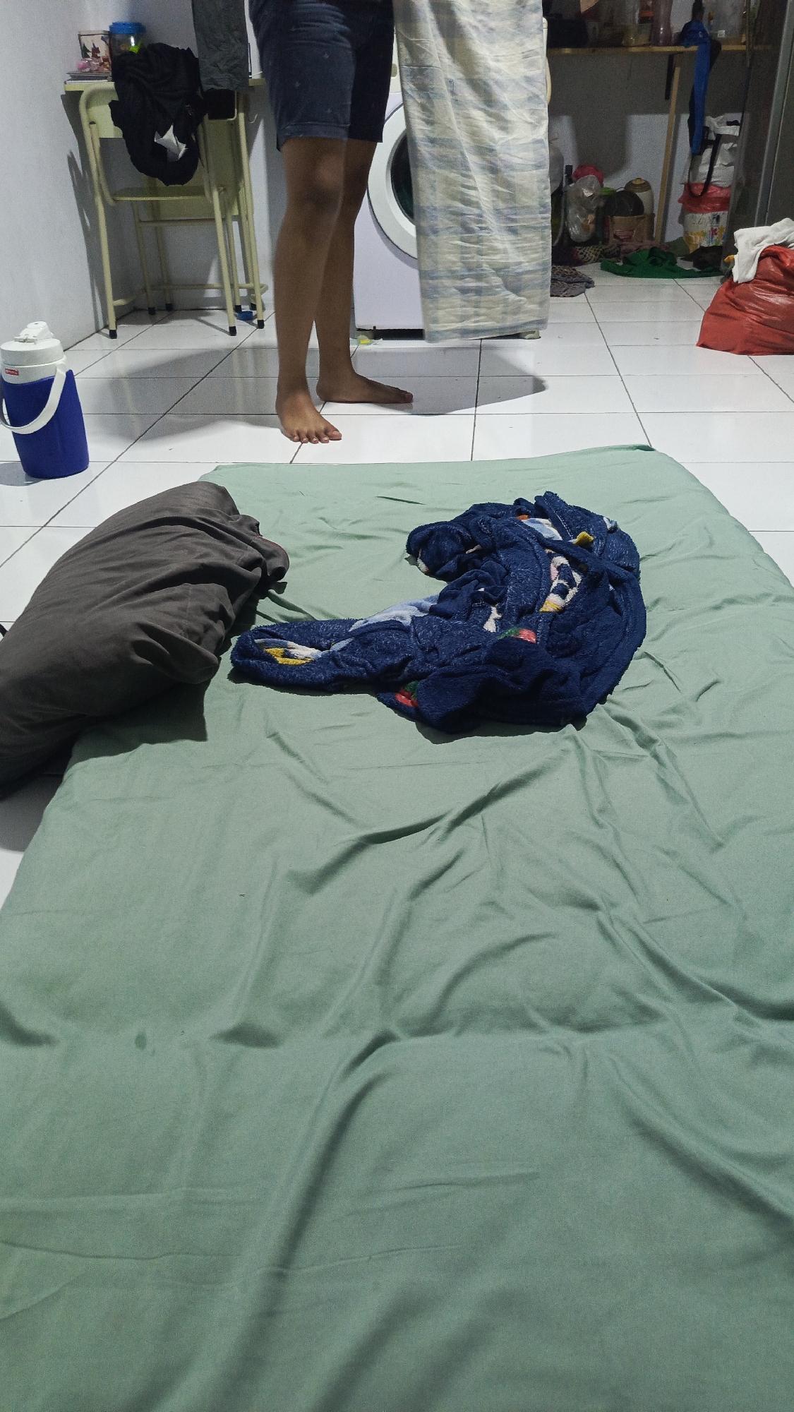 In a room with white tiled flooring, likely in Parung Panjang, Indonesia, a person is standing near a washing machine, holding a piece of patterned fabric. In the foreground, a green mattress is laid out on the floor, with a large gray pillow on the left and a pile of dark blue folded clothes or blankets on the right. A white and blue water jug is visible to the left of the person, and shelves with various items are in the background.