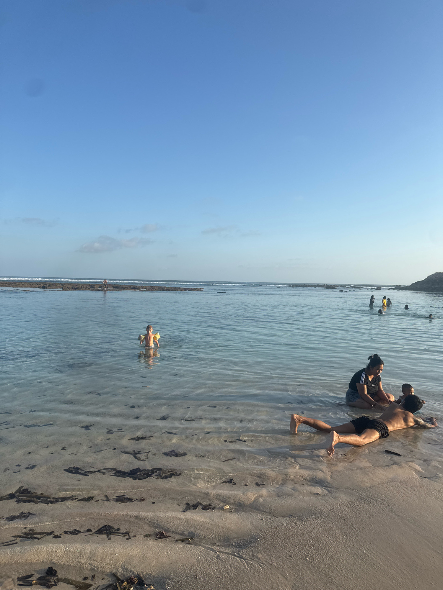 The image depicts a shallow ocean bay with a sandy bottom, located in Kuta Selatan, Indonesia. The sky is clear and blue, with a few faint clouds near the horizon. In the foreground, a man lies face down on the sandy seabed, his legs extended. Beside him, a woman is sitting in the water, with a baby on her lap. Further out in the bay, several other people are visible, some swimming and others standing in the shallow water. A young child with yellow armbands is standing in the water, facing the camera. In the distance, a rocky reef is visible, with waves breaking over it. The overall scene suggests a calm and leisurely day at the beach.