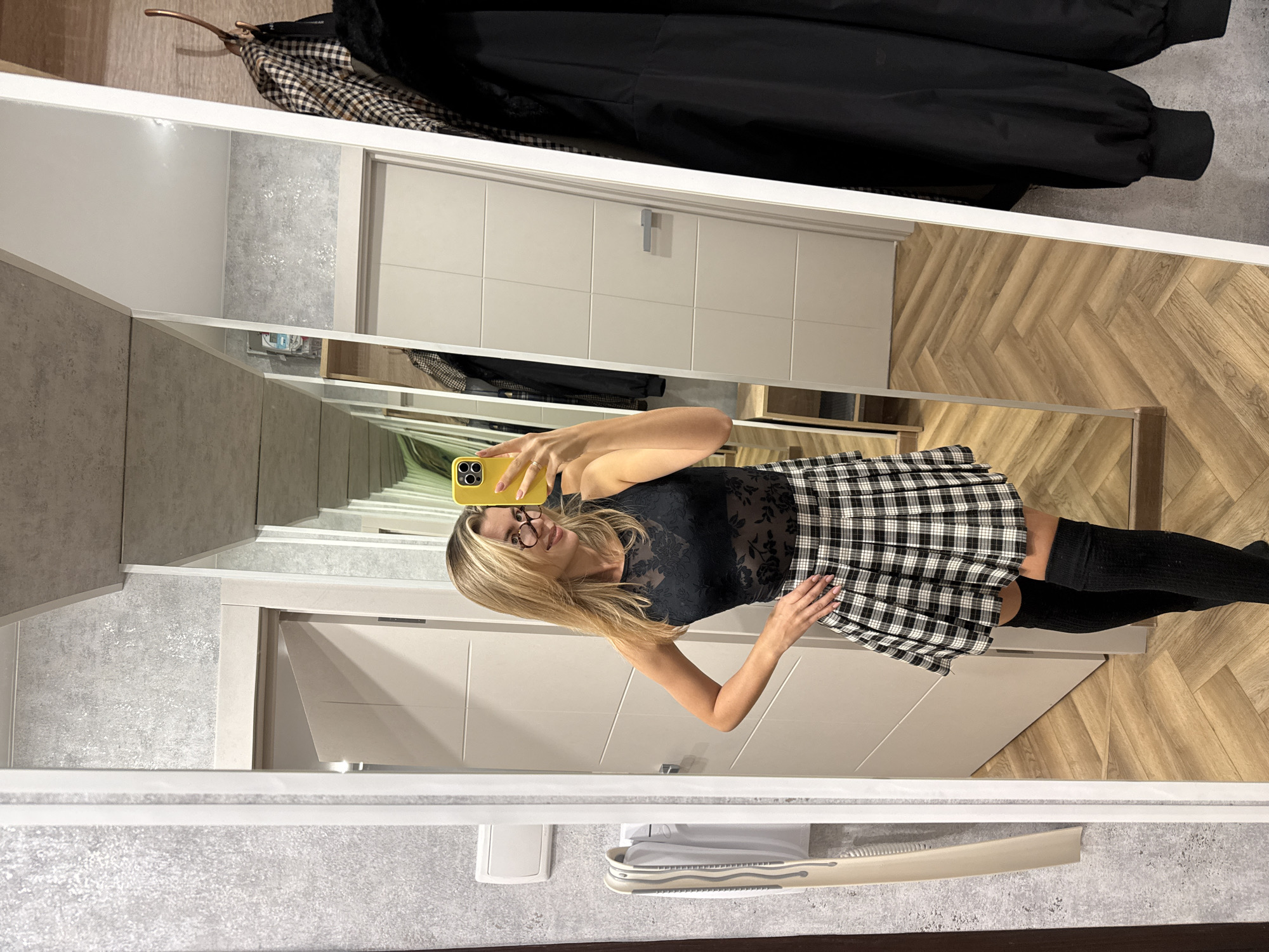 The image, rotated 90 degrees clockwise, captures a young woman taking a mirror selfie indoors. She is seen from the chest up in the reflection, holding a yellow-cased smartphone with a triple camera array to capture her image. She has long, straight blonde hair and is wearing round, clear-framed glasses. Her expression is a subtle smile.
She is dressed in a black sheer lace top with a floral pattern, a black and white plaid pleated mini skirt, and black thigh-high socks. A silver-colored ring is visible on her left hand, which holds the phone, while her right hand rests on her hip.
The setting appears to be a modern interior space, possibly a hallway or entrance. The floor is made of light brown wood planks laid in a herringbone pattern. The walls feature a textured light grey finish, complementing smooth white panels and doors visible in the background. Above the mirror, several items of clothing are hung on a rack, including a black jacket and a black and brown plaid coat. No discernible text is visible in the image.