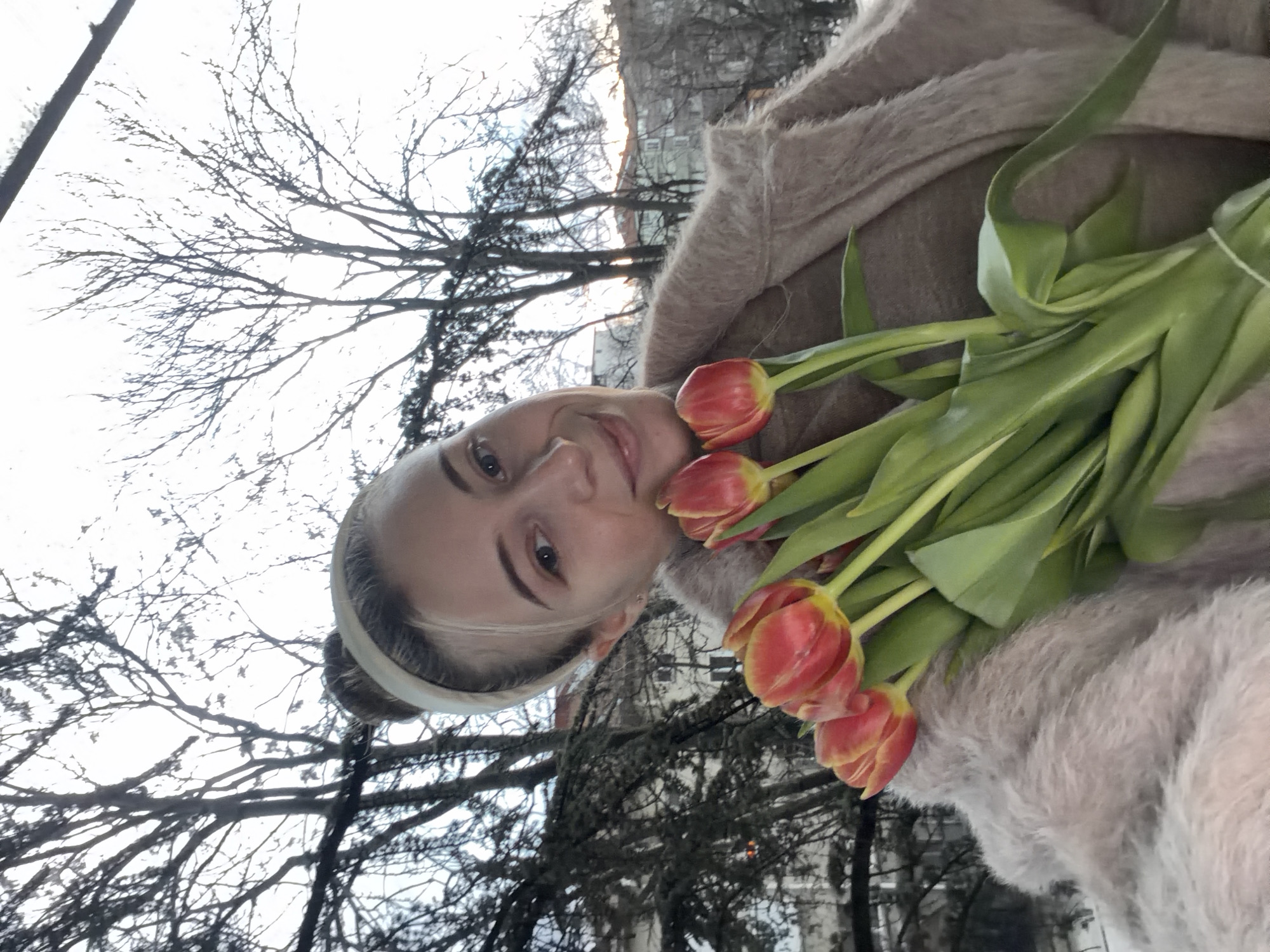 A woman with light hair and a headband is holding a bouquet of red and yellow tulips in an outdoor setting. The background reveals bare tree branches and building facades, suggesting an urban location. The lighting suggests it's either early morning or late afternoon, and given the bare trees, likely the end of winter or early spring. There are no other people visible in the immediate frame, and the focus is on the woman and the flowers. Considering the architecture and the bare trees in conjunction with the tulip bouquet, it is likely that this photo was taken in Plovdiv, Bulgaria in the early Spring.