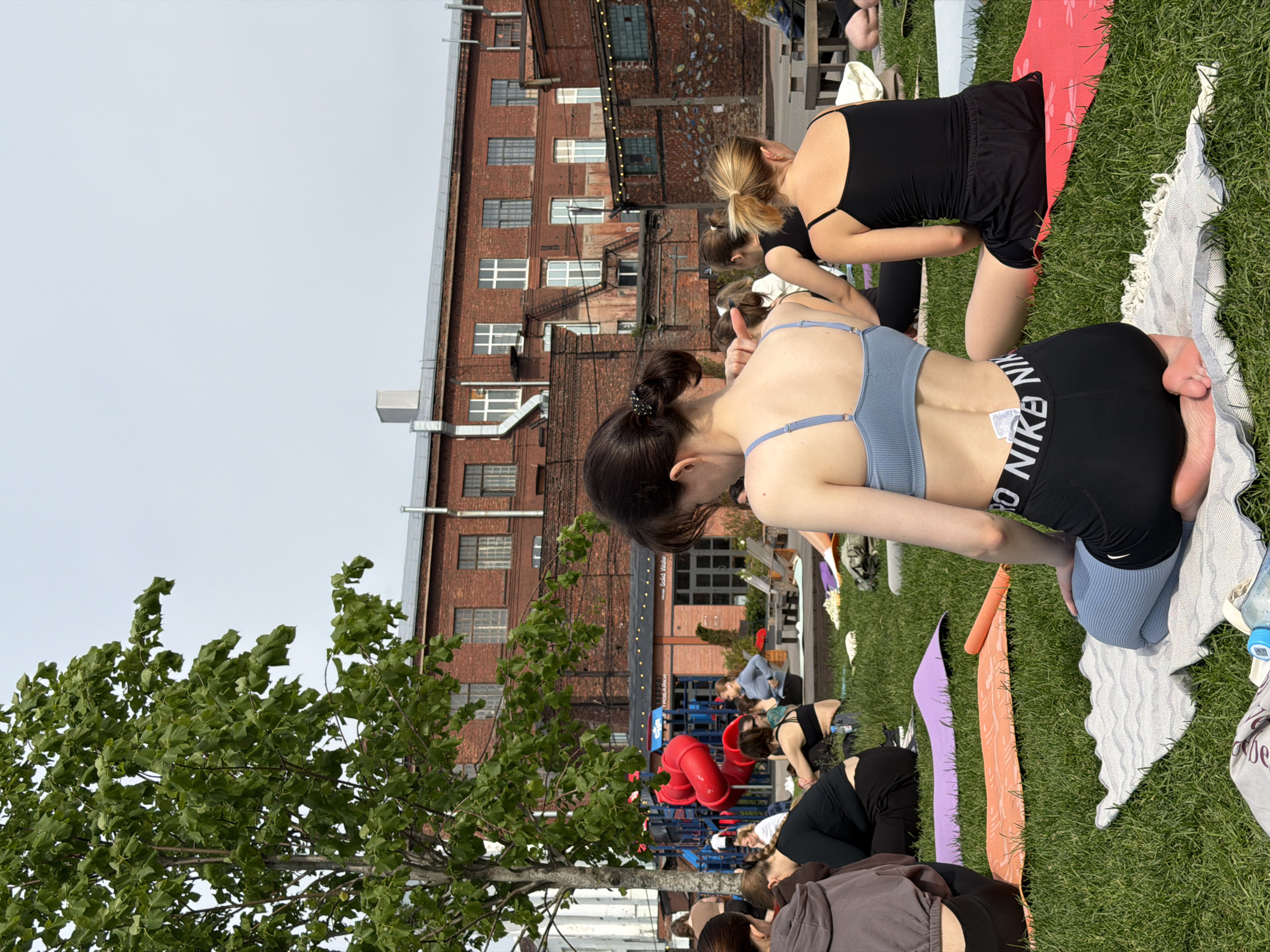 The image captures an outdoor group yoga or fitness session on a grassy lawn at Sevkabel Port in Saint Petersburg, Russia. The sky is overcast, providing diffuse natural lighting.
In the foreground, a young woman with dark hair styled in two small buns kneels on a light grey and white patterned towel, facing away from the viewer. She is wearing a light blue ribbed sports bra and black athletic shorts with "NIKE" in white lettering on the waistband. Her hands rest on her thighs, and her bare feet are visible beneath her. To her immediate right, another woman sits on a red yoga mat, dressed in a black tank top and black shorts. Scattered across the green grass, multiple other individuals, primarily women, are seated or kneeling on various colored yoga mats, including orange and purple, participating in the outdoor activity. A partially visible blue-capped water bottle lies near the foreground towel.
The background is dominated by multi-story red brick industrial-style buildings, characteristic of the repurposed Sevkabel Port. These structures feature numerous rectangular windows, some with white frames, and external metal fire escapes. A large tree with vibrant green leaves occupies the left side of the frame, partially obscuring a white building. Below the tree, a children's playground with distinctive bright red tubular slides is visible on a light-colored surface. Signs on the background buildings include partial text such as "Solid Waste" and Russian words "УПРАЖНЕНИЯ" and "ПРОДАЖИ," indicating commercial or retail establishments within the complex. Wooden outdoor seating and string lights are also visible on the building facades in the midground.