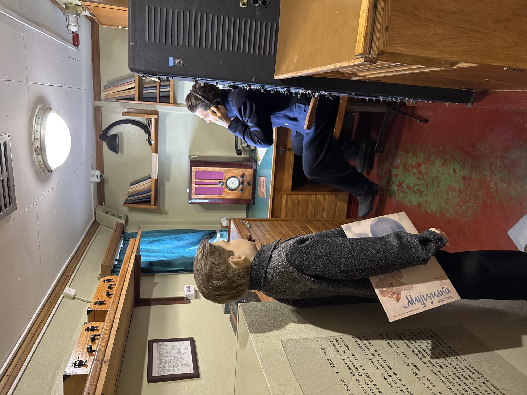 The image shows a diorama set within what appears to be a museum exhibit, possibly depicting a radio operator's station in Saint Petersburg, Russia, given the inscription on the wall. In the foreground, a mannequin of a young boy stands facing left, holding a magazine with Cyrillic text, likely titled "Murzilka." Behind him, another mannequin, dressed in a dark blue suit, is seated at a wooden desk, wearing headphones and appearing to operate a radio. The room is filled with vintage equipment, including a large, dark speaker system to the right, a radio set on the desk, and shelves lined with books and what look like early communication devices. A desk lamp illuminates the workspace. The walls are a pale green, and a bright overhead light fixture is visible on the ceiling, which also has visible ductwork. A red and green stained floor suggests an older industrial or maritime setting. The wall to the left features a framed schematic and further Cyrillic inscriptions.