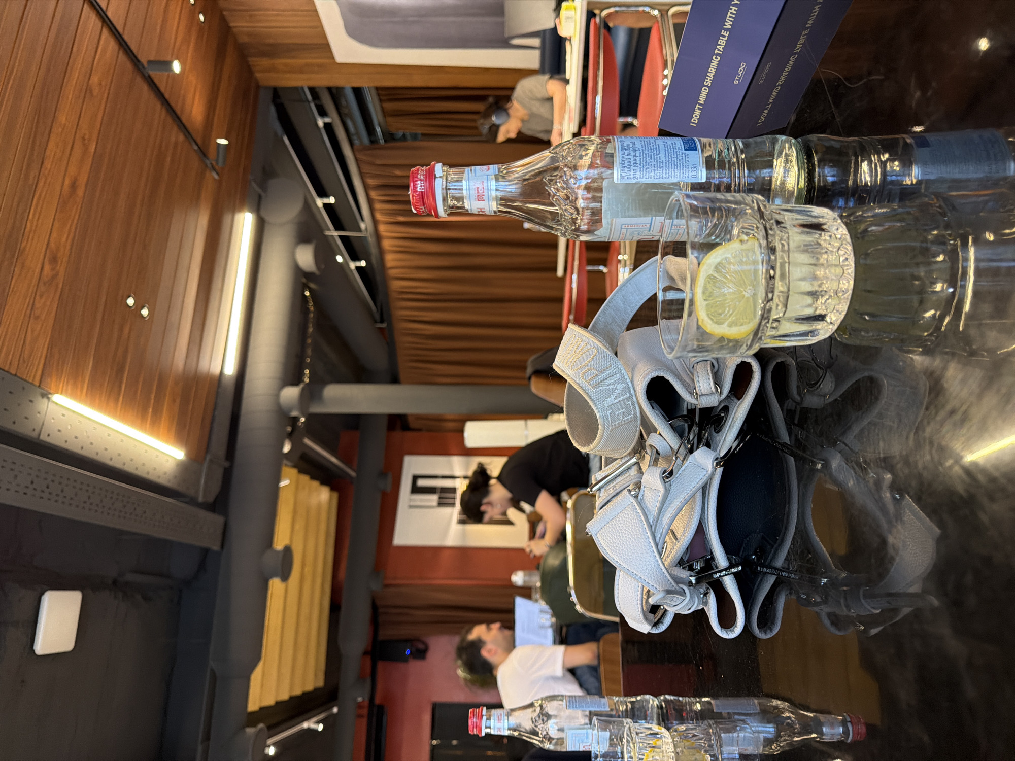 A brightly lit interior space, likely a restaurant or cafe, reveals a communal table setting. On the table are two glass water bottles, one with a red cap, labeled with Armenian text that translates to "Crystal Spring Natural Drinking Water, Non-Carbonated" alongside a glass of water with a lemon slice. A white shoulder bag sits next to the beverages, reflecting on the shiny table surface. A book titled "I Don't Mind Sharing Table With You" is also present. In the background, people are seated at the table, engaged in activities like eating and using computers. Based on the bottle label and the interior design, the location is possibly in Yerevan, Armenia.