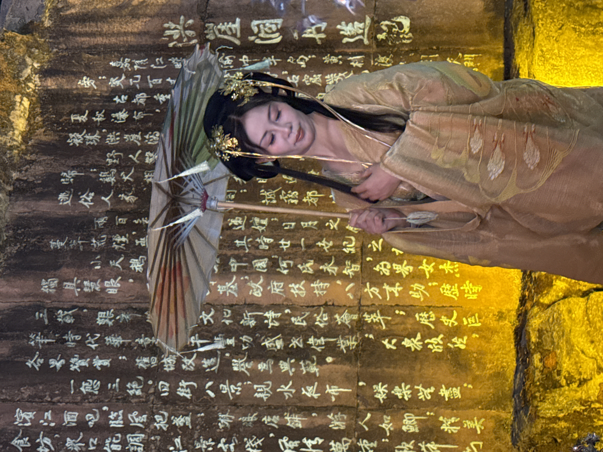 An East Asian woman dressed in a traditional gold-colored robe and ornate gold hairpieces stands in front of a stone wall covered in Chinese calligraphy. She is holding a decorative umbrella. The woman is looking down and to her right, with her hand resting on the umbrella. The stone wall is illuminated by yellow light, highlighting the white inscriptions. The scene appears to be outdoors, possibly in a historical or cultural site within Yuzhong District, China, suggested by the style of dress and the presence of calligraphy on the stone.