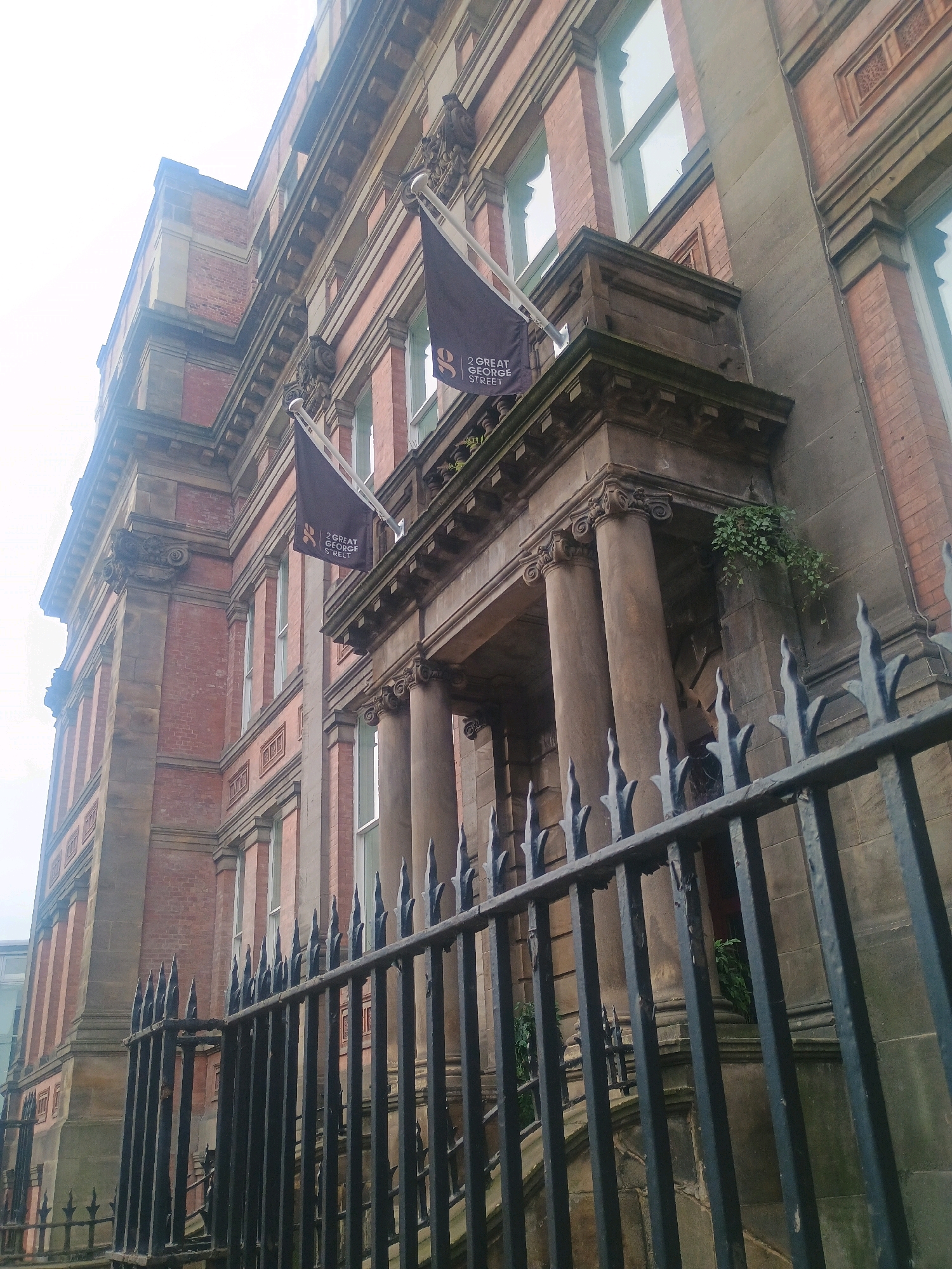 This is a low-angle, full shot of a red brick building facade with ornate architectural details. The building has multiple stories with numerous windows. Two dark flags with white lettering are visible, each bearing the text "2 GREAT GEORGE STREET" and a stylized "G" logo. The flags are attached to poles extending from the building. A black wrought-iron fence with pointed finials runs across the foreground. The sky is overcast and bright. The building appears to be in an urban setting.