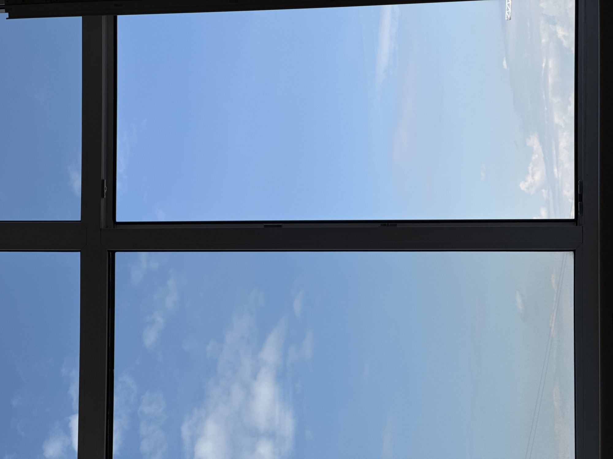 The image displays a view through a large window, divided into multiple panes by dark gray or black frames. The dominant subject is the clear blue sky, visible through all the window sections. The top section of the window appears slightly open, or its frame is angled, creating a thinner horizontal bar.
Through the windowpanes, the sky is a bright, light blue, indicative of daytime. Some white, wispy clouds are scattered across the sky, particularly noticeable in the lower right and upper right sections, and faintly in the lower left. The lighting suggests a bright, sunny day.
There is no discernible activity or event taking place, as the image captures a static outdoor scene from an indoor perspective. No people or objects are visible beyond the sky.
A notable detail is a faint, inverted reflection or marking in the top right pane, which appears to be some kind of text or alphanumeric sequence, possibly "03RN" or similar, although it is distorted and difficult to read precisely. The reflection suggests the window material is slightly reflective or there is something behind the camera reflecting onto the glass. The overall scene conveys a calm, uneventful atmosphere.