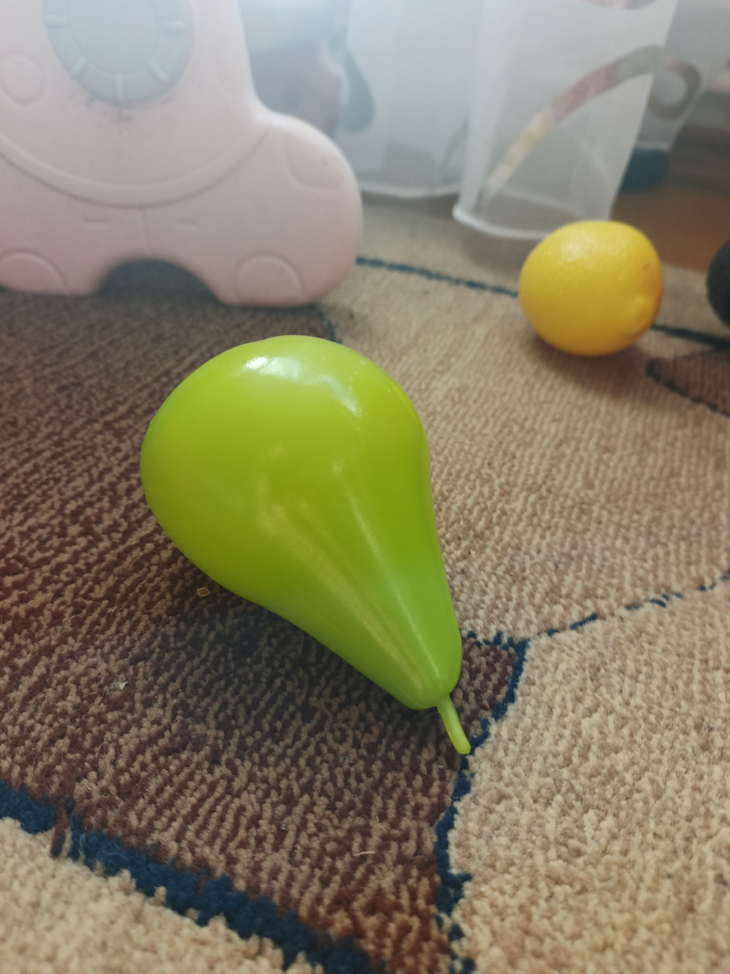 This close-up, eye-level shot features a collection of colorful plastic toys on a patterned carpet. In the foreground, prominently positioned slightly left of center, is a bright, glossy green toy pear with a small stem. To its right, further into the background, lies a round, yellow plastic toy lemon. On the far left, a blurred section of a pink plastic toy, possibly a children's ride-on toy like a rocking horse, is visible.
The setting appears to be indoors, likely a home, with the primary surface being a textured carpet featuring patterns in shades of brown, beige, and dark blue. The lighting suggests daytime, with soft ambient light creating subtle reflections on the shiny surfaces of the toys. No specific activity is depicted, as the toys are static. There is no visible text or discernable emotions in the scene. The background is softly blurred, indicating a shallow depth of field, with some indistinct white elements possibly belonging to furniture or curtains.