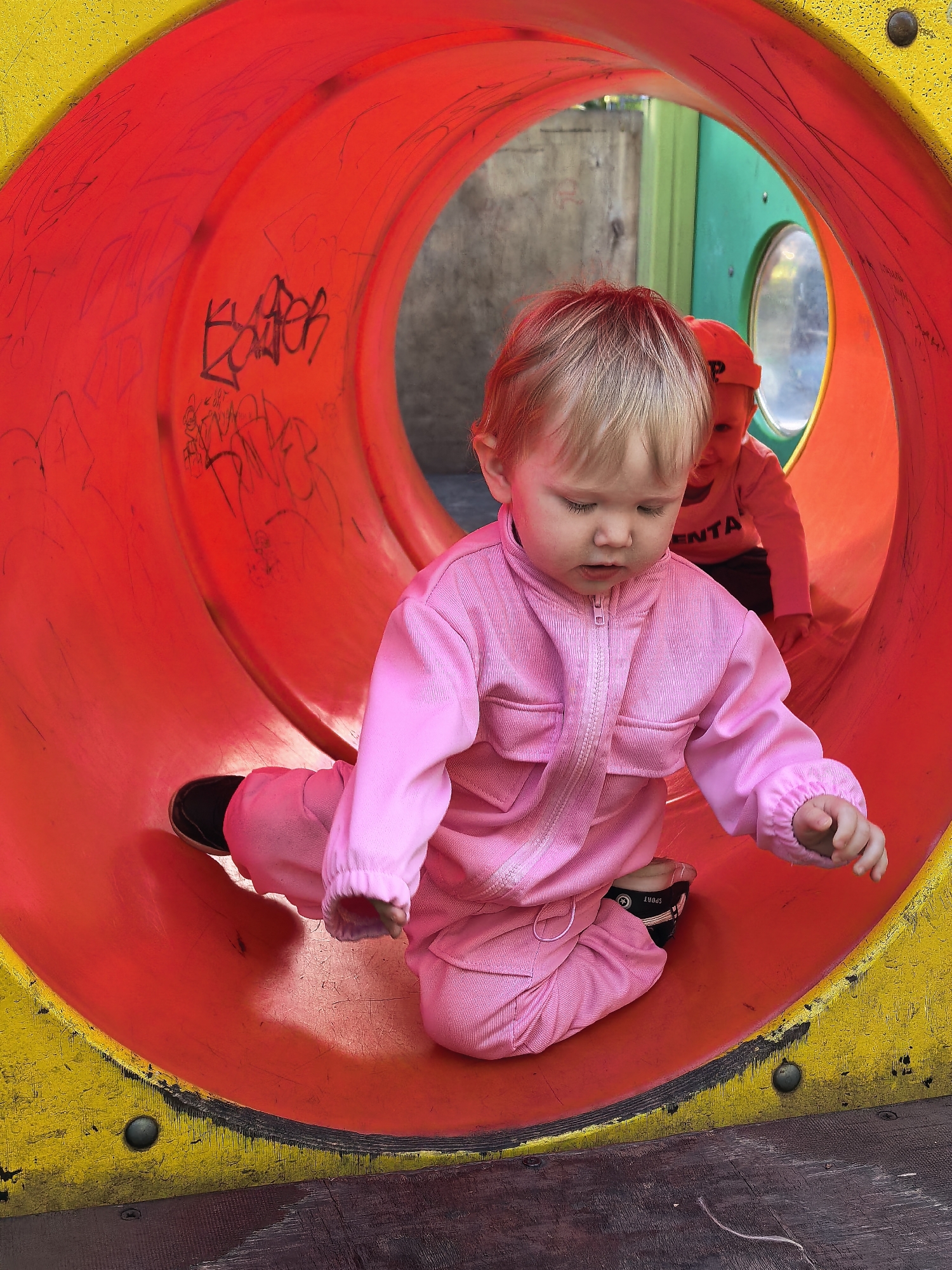 This is a photograph of two young children inside a red and yellow tubular playground tunnel. The child in the foreground, with blonde hair, is wearing a pink jumpsuit and black shoes with white accents. The child's eyes are closed, and their head is tilted downwards, suggesting they are focused on something in front of them or resting. Their right hand is extended slightly, with fingers open. Another child, partially visible in the background, is wearing a red shirt and a red cap, and appears to be looking towards the front child. The tunnel has graffiti scribbled on the interior red surface. The playground structure appears to be made of durable plastic and metal. The ground beneath the tunnel is a dark, textured material. The overall environment suggests an outdoor urban playground setting. The specific location is not determinable from the image alone; there is no indication of Sterlitamak, Russia.