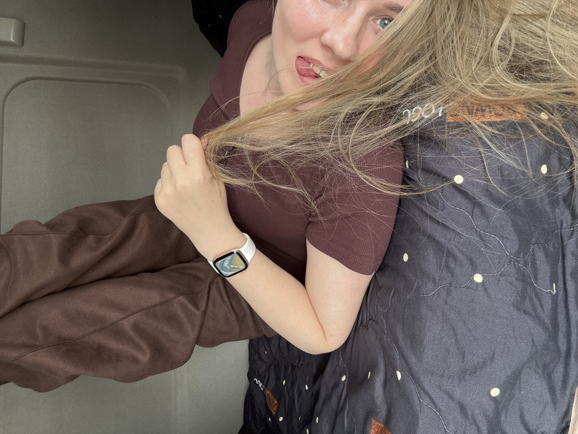 A young woman with fair skin and blonde hair, wearing a brown short-sleeved shirt and brown sweatpants, sits in what appears to be a vehicle or small enclosed space with gray walls. She is positioned on a surface covered with a dark fabric patterned with light-colored dots. She holds a section of her hair in her left hand and is sticking out her tongue. A white smartwatch is visible on her left wrist. The context lacks specific elements to definitively place the image in Izhevsk, Russia.