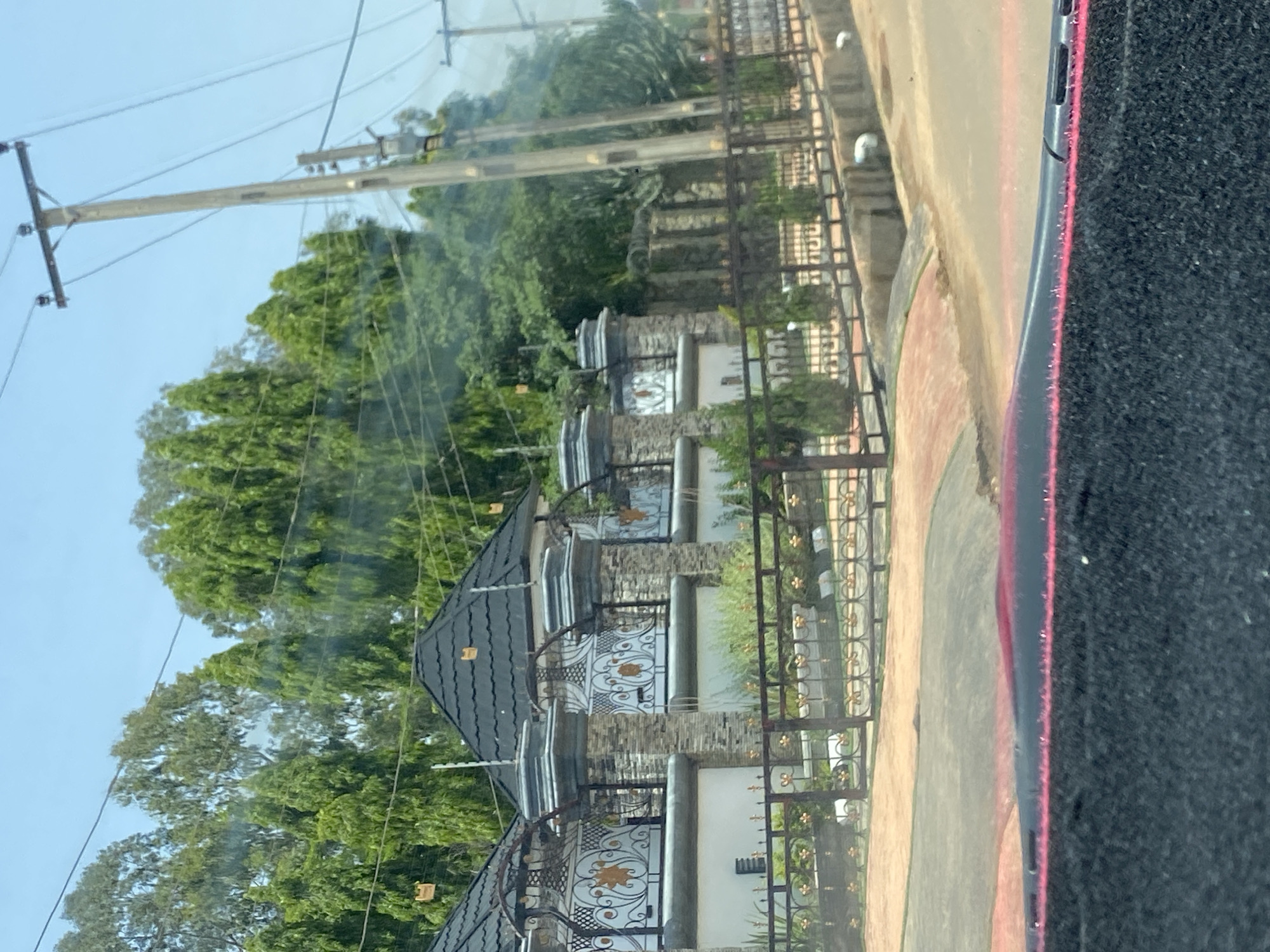 The image depicts a view from a vehicle in the city of Gura Top, Nigeria. In the foreground, the dashboard or interior of the vehicle is visible, with a dark, textured material and a strip of red light. Beyond the vehicle's interior, a street or road is visible with a paved surface and a curb. On the right side of the road, a concrete drainage channel is present.
Further down the street, a large, ornate fence made of metal with gold accents encloses a property. Behind the fence, a building with a dark, tiled roof and stone pillars is partially visible. The building features decorative white panels with orange floral motifs. Greenery, including trees and shrubs, surrounds the property and extends into the background. Several utility poles with electrical wires are also present along the roadside. The sky is clear and blue, indicating a sunny day. There are no people or animals visible in the image. The overall scene suggests a residential or commercial area with well-maintained landscaping and architecture.