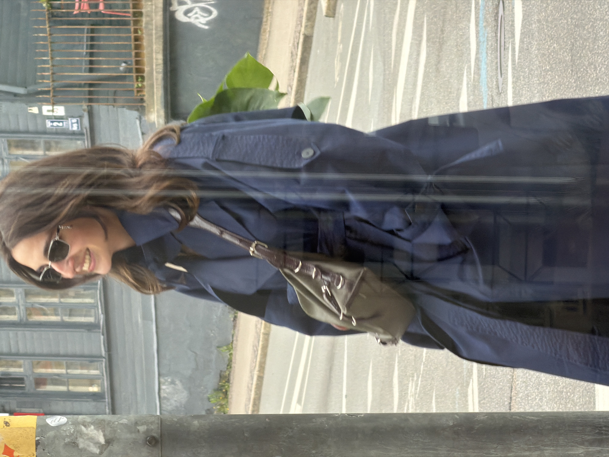 Captured through a possible reflective surface, the image shows a woman in sunglasses and a dark blue trench coat, carrying a shoulder bag, likely on a street in Riga, Latvia. Behind her, a building with many windows and a gray facade is visible. A portion of a road runs vertically in the background. A section of a pole or structure is in the immediate foreground.