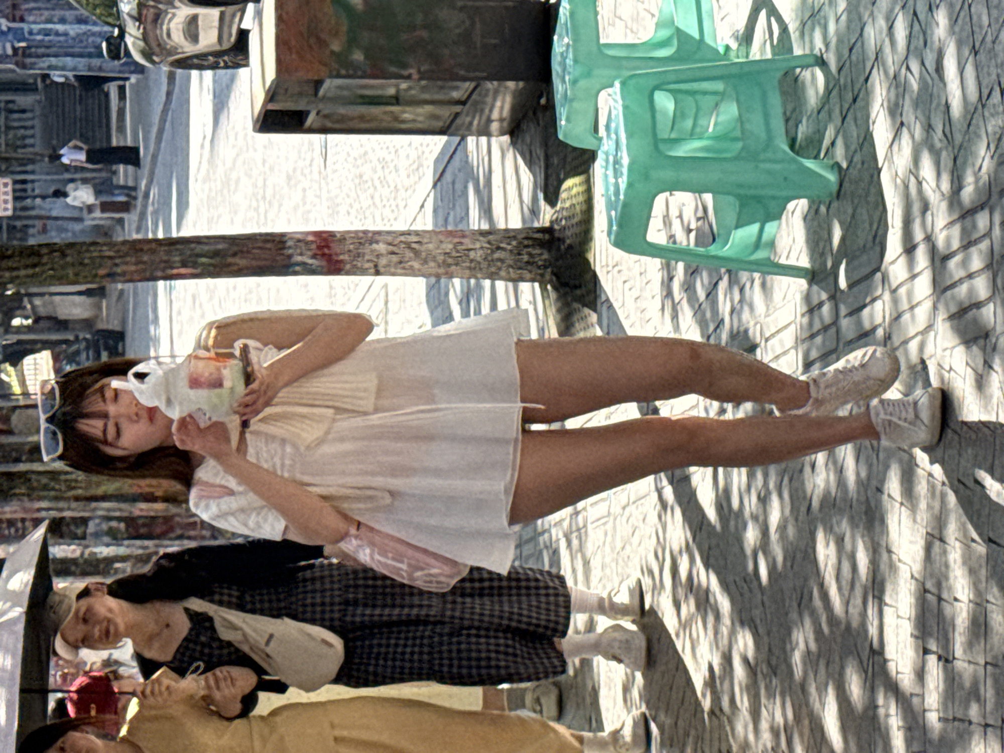 The image depicts a street scene in Jiulongpo District, China. In the foreground, a young woman in a white dress, sunglasses, and white sneakers drinks from a beverage container. Her legs are exposed, and she is standing on a patterned brick surface partially shaded by trees. Behind her and slightly to the left, other people are visible, including a woman with a checkered dress and a woman wearing a yellow top. A black umbrella covers at least one of the people. Behind the woman in the white dress, there are two light green plastic chairs stacked on top of each other and a tall trash receptacle. Further in the background, a building and people walking are visible. The lighting suggests it is a sunny day.