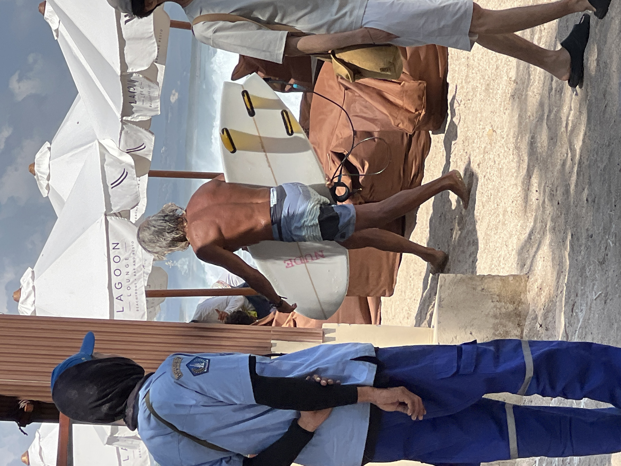 The image depicts a beach scene in Kuta, Indonesia, likely at a beachfront establishment. In the foreground, a person wearing a blue uniform with reflective stripes on the pants and a black head covering with a blue cap is standing and facing away from the camera. This individual appears to be an employee or staff member. To the right, a shirtless person with long grey hair is walking away from the camera, carrying a white surfboard with yellow fins. They are wearing patterned swim trunks. In the background, several large white umbrellas with "Lagoon Lounge" branding are visible, suggesting this is an area for patrons of a lounge or restaurant. The ground is sandy, and the ocean with moderate waves can be seen in the distance. Another person, partially visible on the far right, is wearing a light-colored t-shirt and white shorts, and appears to be walking away from the viewer. Brown beanbag-like cushions are scattered on the sand. The sky is partly cloudy.
