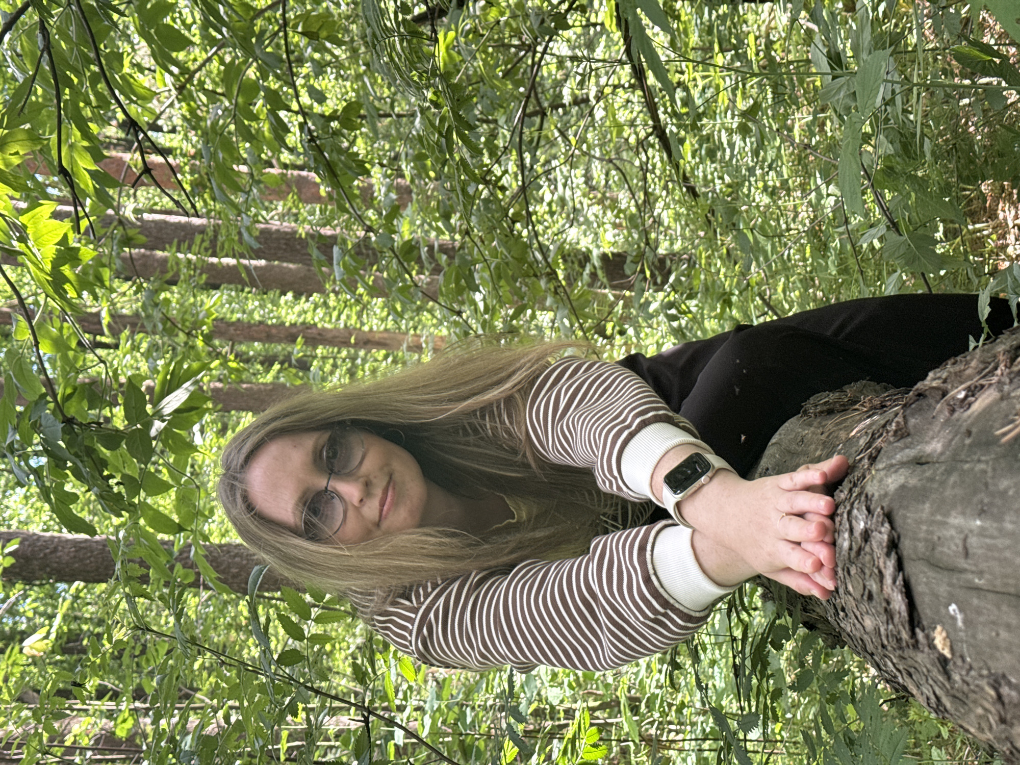 A woman with blonde hair and glasses is sitting on a log in a forest. She is wearing a striped shirt and dark pants. The trees are tall and green, with sunlight filtering through the leaves. The woman is looking at the camera and smiling. The log is thick and rough, with bark visible. Her hands are resting on the log, and she is wearing a watch on her left wrist. The forest is dense and full of foliage, creating a natural and serene environment. The scene is set in Chaikovsky, Russia.