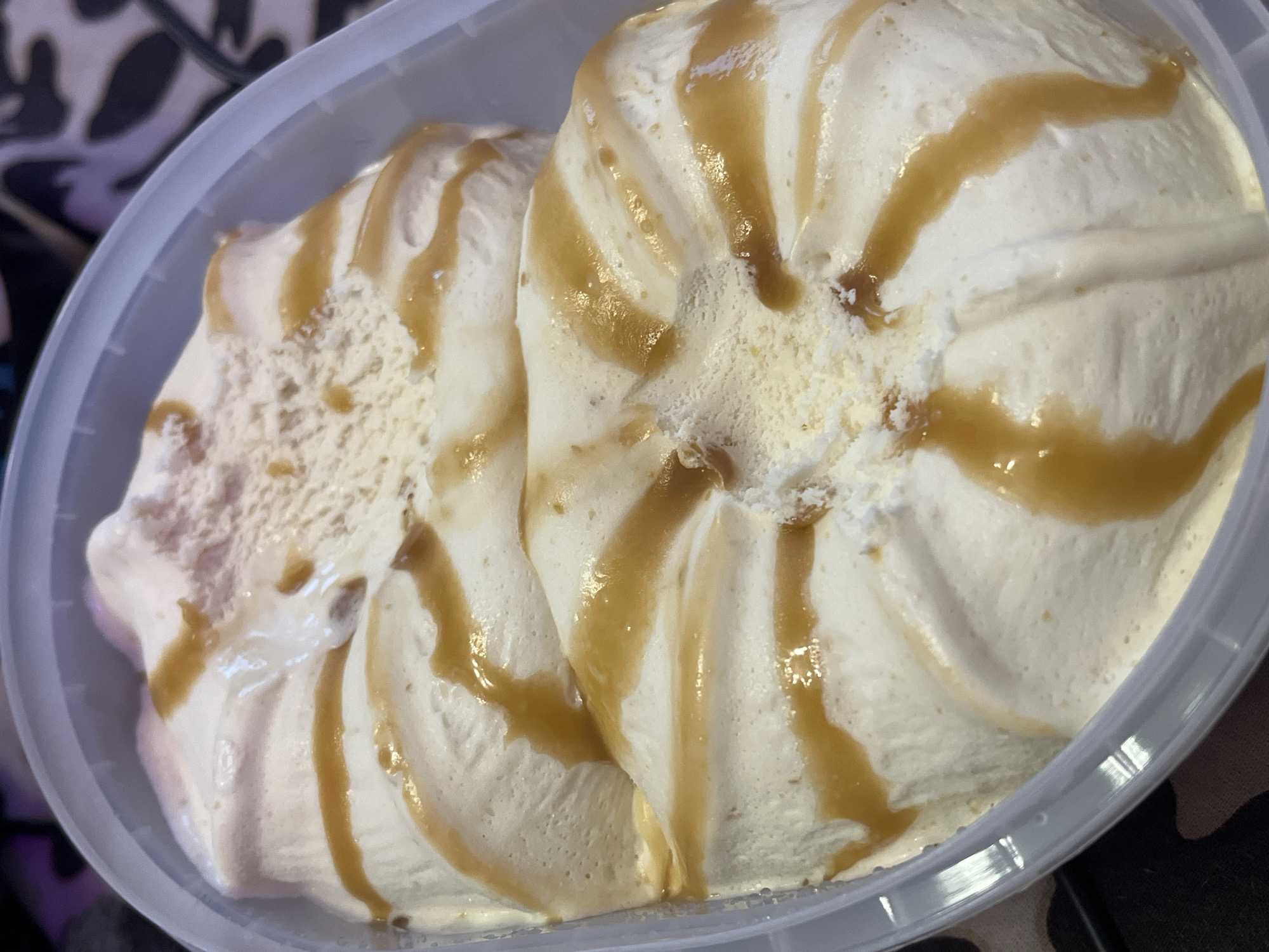 A container of ice cream with caramel swirl is shown from an overhead perspective. The ice cream is white and creamy, with visible texture. Caramel sauce is drizzled across the top in an irregular, circular pattern. The container appears to be a translucent plastic tub. The background is out of focus but appears to be a patterned surface. There is no evidence in the image to indicate the location as Saint Petersburg, Russia.