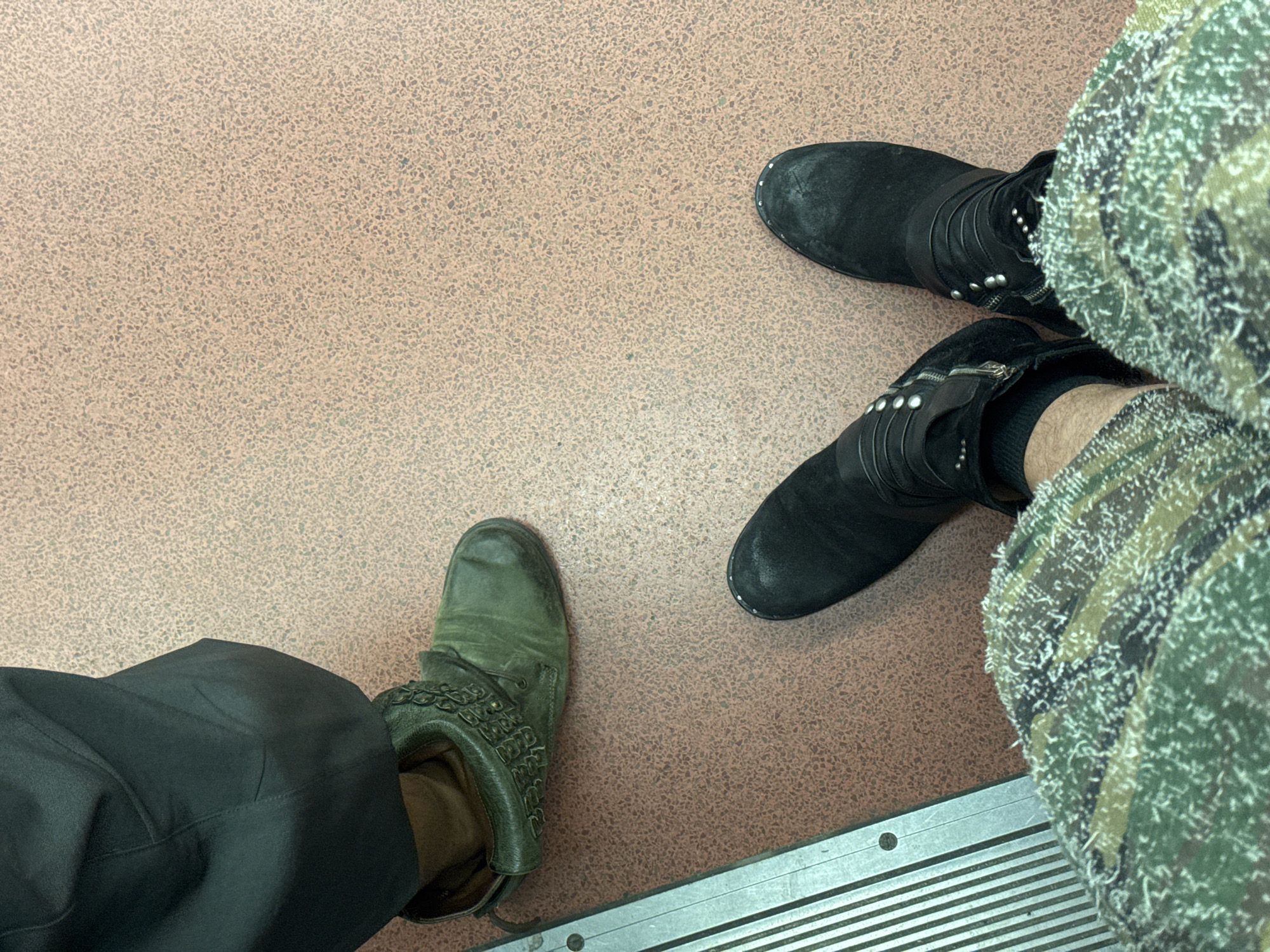 The image shows the lower bodies and feet of two individuals on what appears to be the floor of a public transport vehicle, possibly a train or subway car. The floor has a reddish-tan speckled pattern. On the left, a person wearing dark-colored trousers and green leather boots with decorative chain-like details. On the right, another person is wearing camouflage patterned trousers and black suede boots adorned with studs and zippers. A metal strip with ridges is visible in the bottom right corner. While the scene evokes the atmosphere of a Parisian subway, there is no definitive evidence within the image to confirm this location.